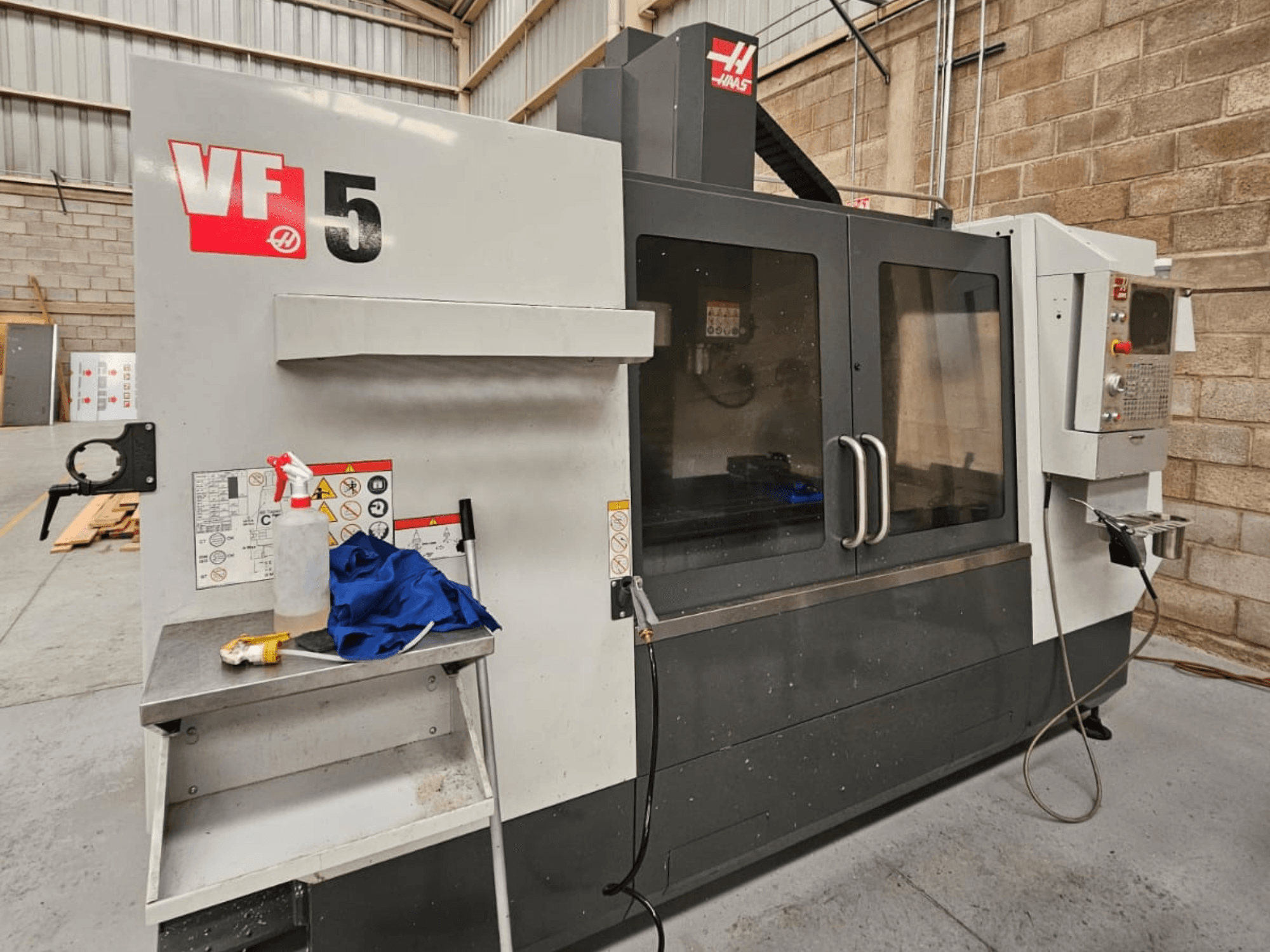 Haas VF-5 CNC glodalica s prednje strane, s upravljačkom pločom, bocom rashladne tekućine, plavom krpom i sigurnosnim uputama.