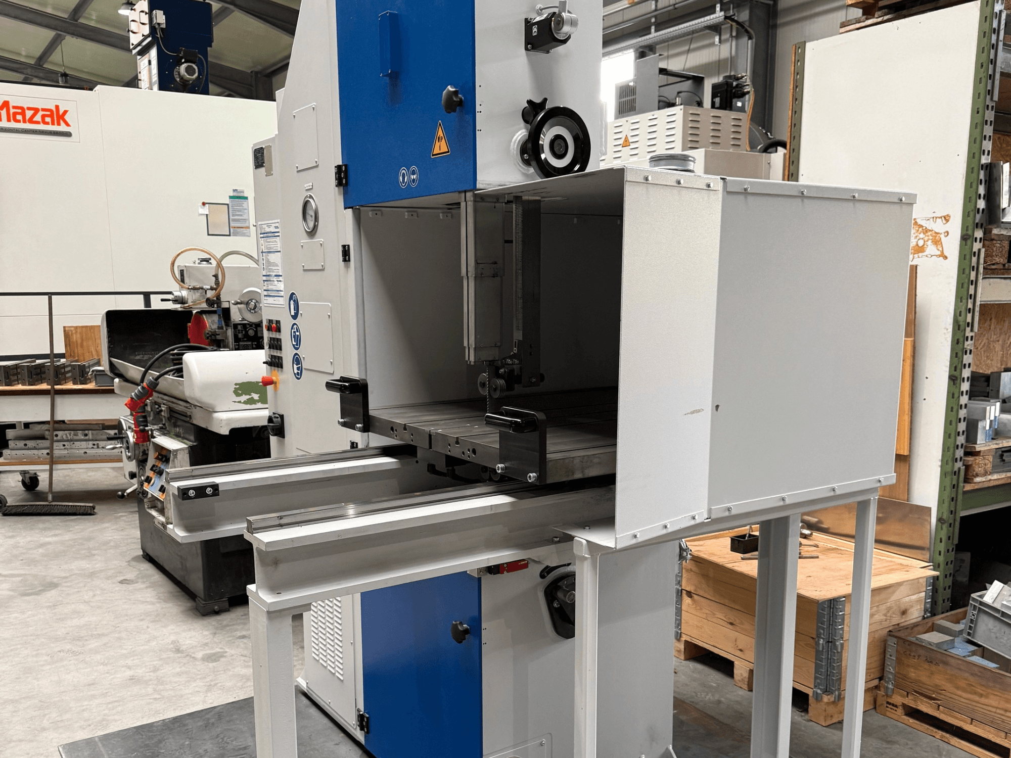 Mazakov industrijski stroj gledan sprijeda, s plavom upravljačkom pločom, metalnim stolom i sigurnosnim upozorenjima.