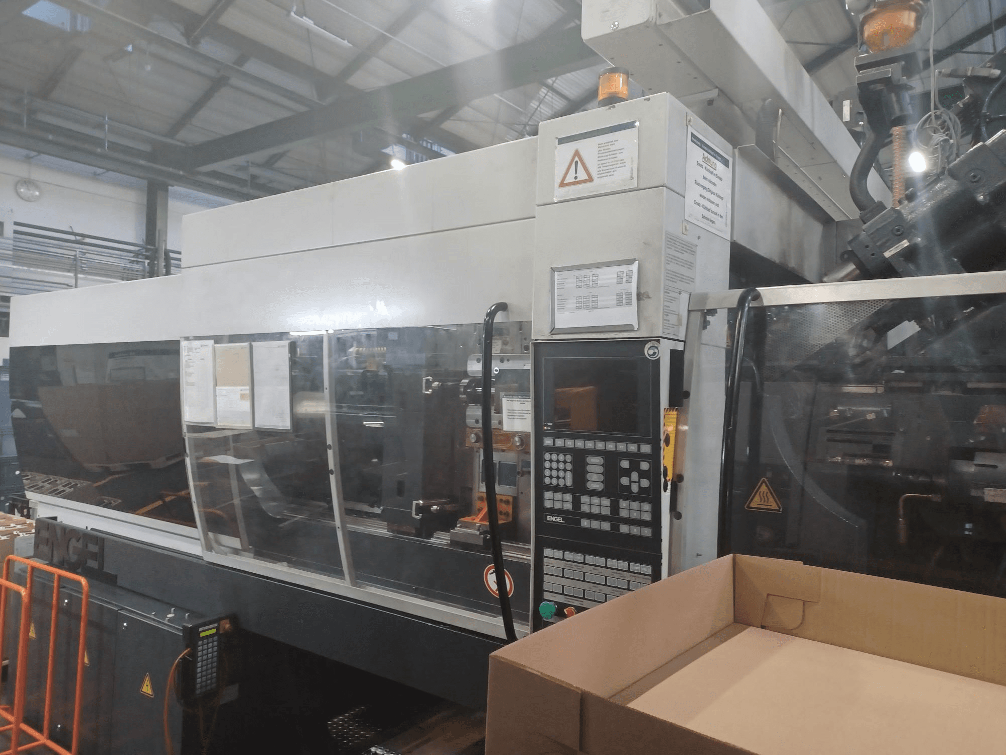 Engel stroj za brizganje plastike, prednji pogled, s upravljačkom pločom, sigurnosnim naljepnicama i praznom kutijom u blizini.