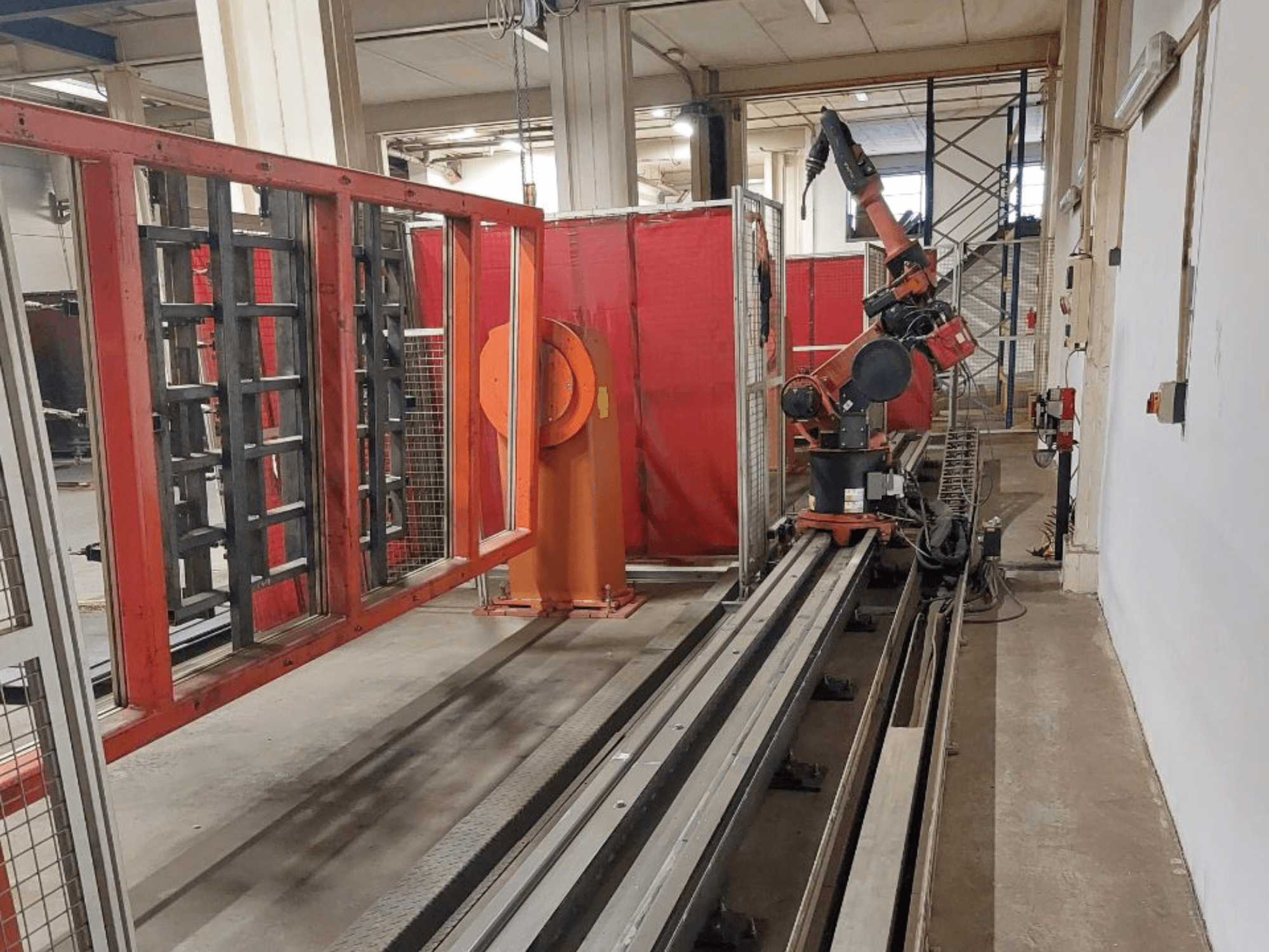 Industrijska robotska ruka narančaste i crne boje postavljena na metalnim tračnicama, okružena crvenim zaštitnim barijerama, pogled sprijeda.