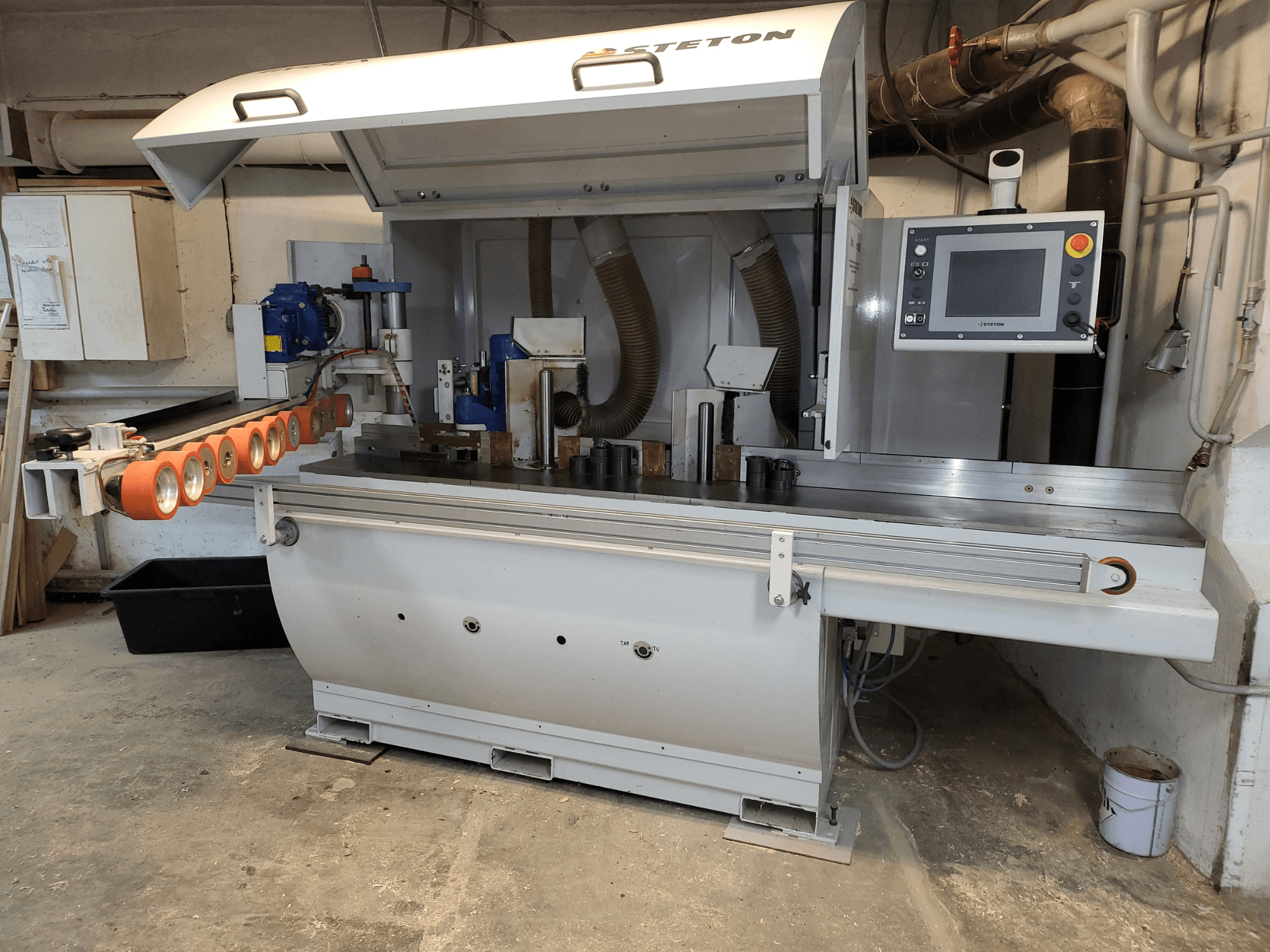 Prednji pogled na Stettonov stroj za obradu drveta s upravljačkom pločom, dovodnim cijevima i raznim drvenim komadima na stolu.