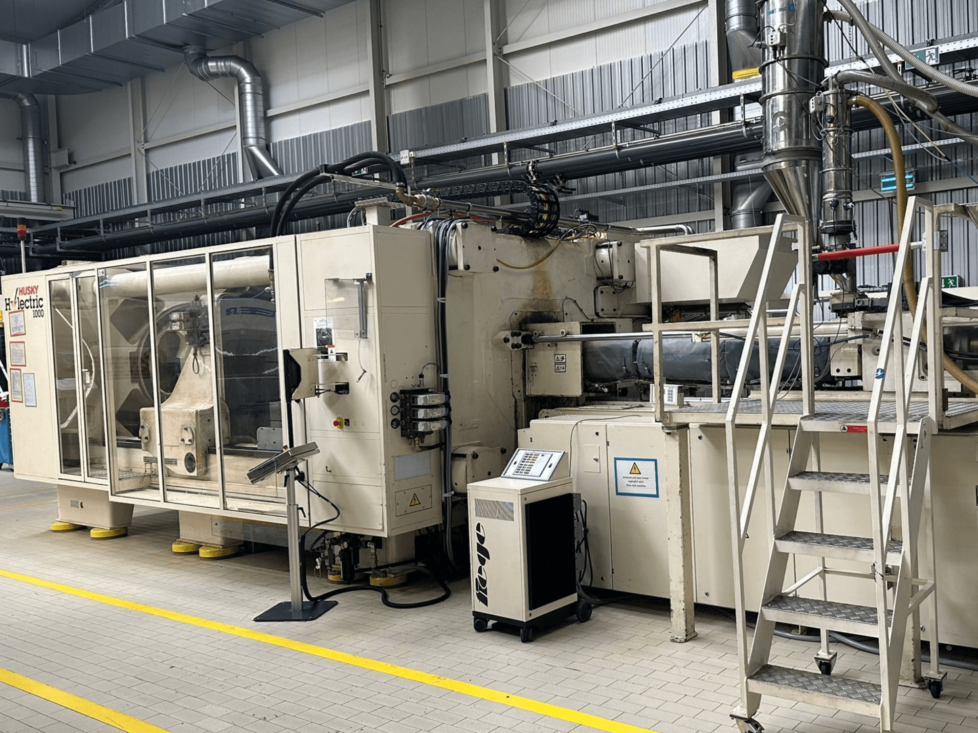 Prednji pogled na električni stroj za brizganje plastike Husky Hylelectric H1000 RS95/80 u tvorničkom okruženju, s upravljačkom pločom i stepenicama.