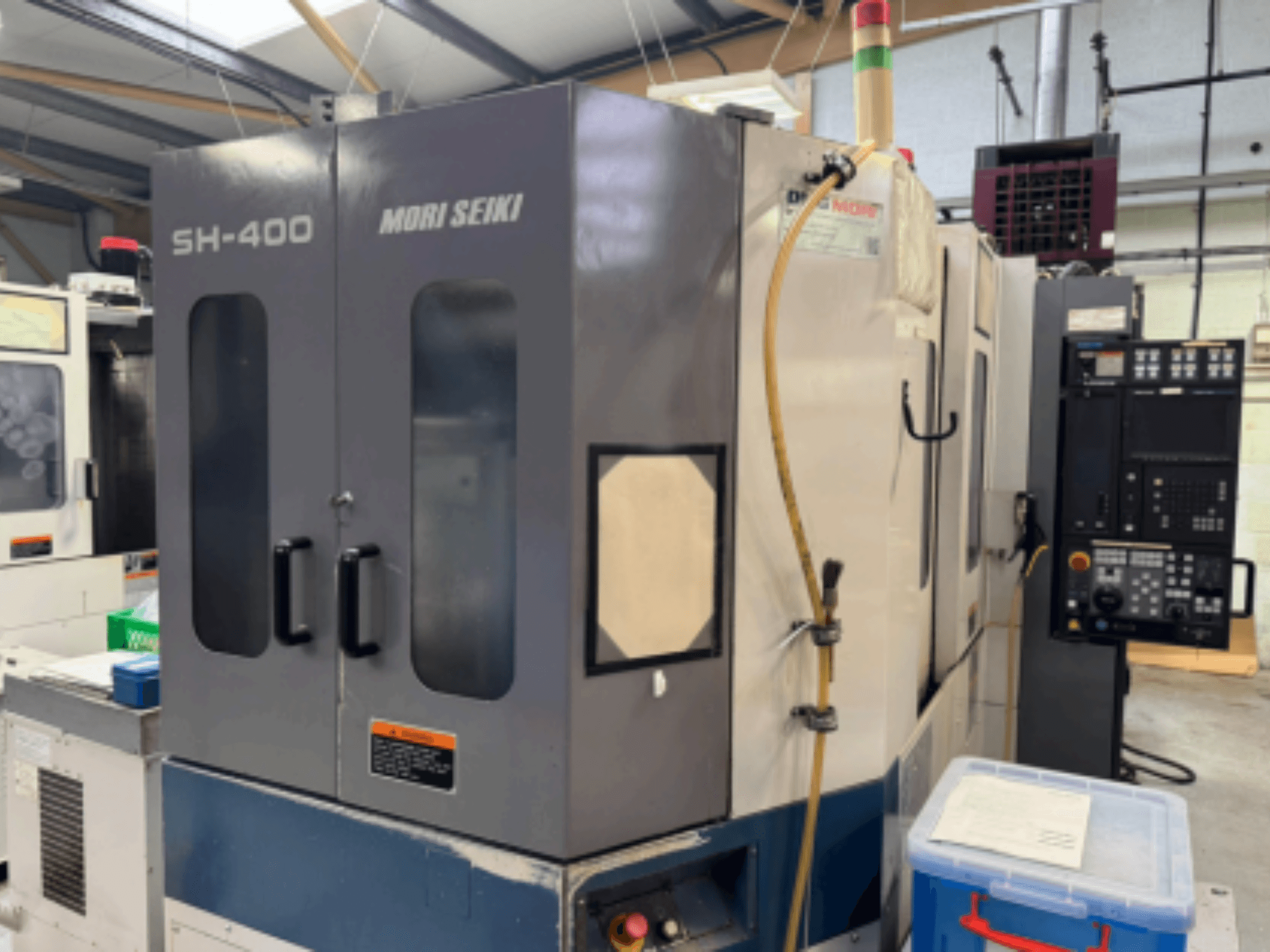 CNC stroj Mori Seiki SH-400, prednji pogled, s dvostrukim vratima, upravljačkom pločom i priključnim crijevom. Plavi spremnik za pohranu u blizini.