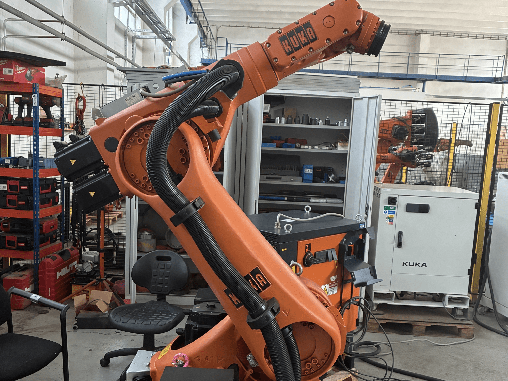Robotska ruka KUKA KR30-3 u industrijskom okruženju, bočni pogled s kabelima i upravljačkim elementima, s alatima i policama u pozadini.