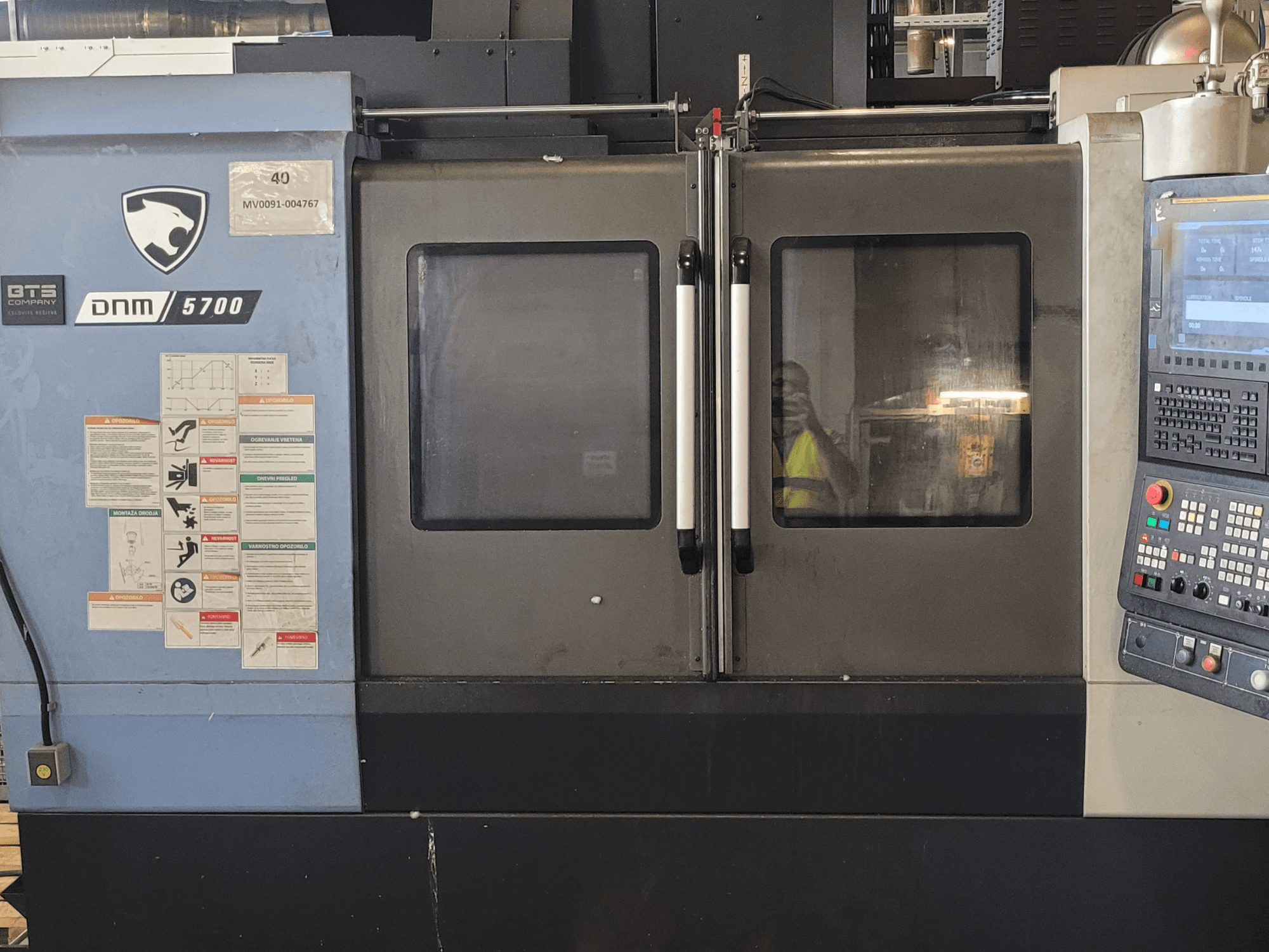 Prednji pogled na vertikalni obradni centar DOOSAN DNM 5700 s upravljačkom pločom, sigurnosnim naljepnicama i blago odškrinutim vratima.