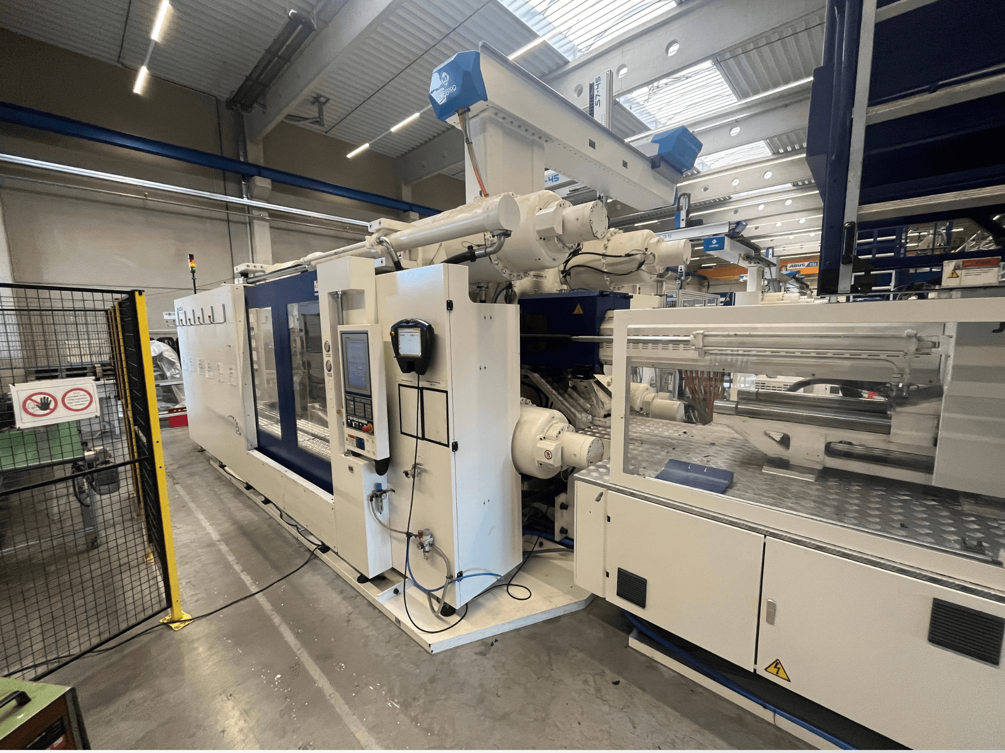 Stroj za brizganje plastike u bijeloj i plavoj boji, prednji pogled, s upravljačkom pločom, robotskom rukom i sigurnosnim znakovima.