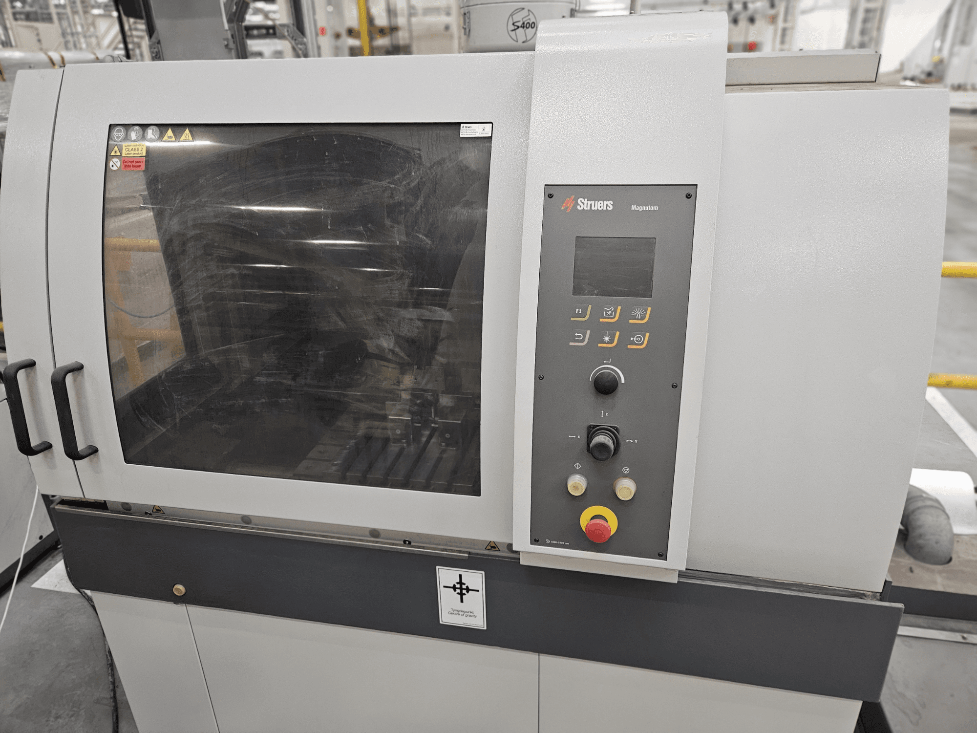 Prednji pogled na Struers Magpentor stroj s upravljačkom pločom, gumbima i velikim prozorom za pregled. Vidljive tehničke oznake.
