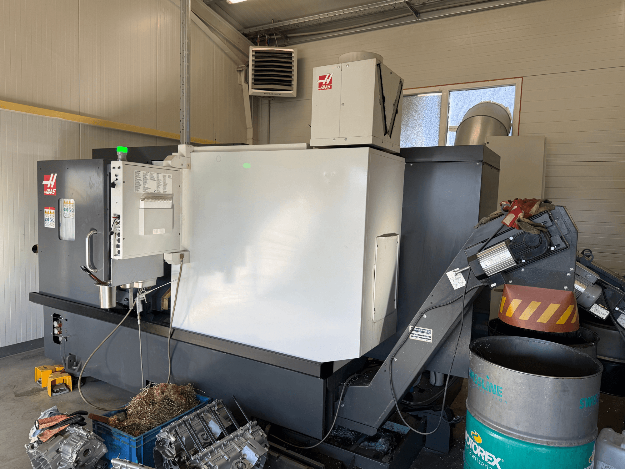 Haas CNC stroj s upravljačkom pločom, bočni pogled; vidljive cijevi za rashladnu tekućinu i ulje; metalni otpad u plavoj kanti; industrijsko okruženje.