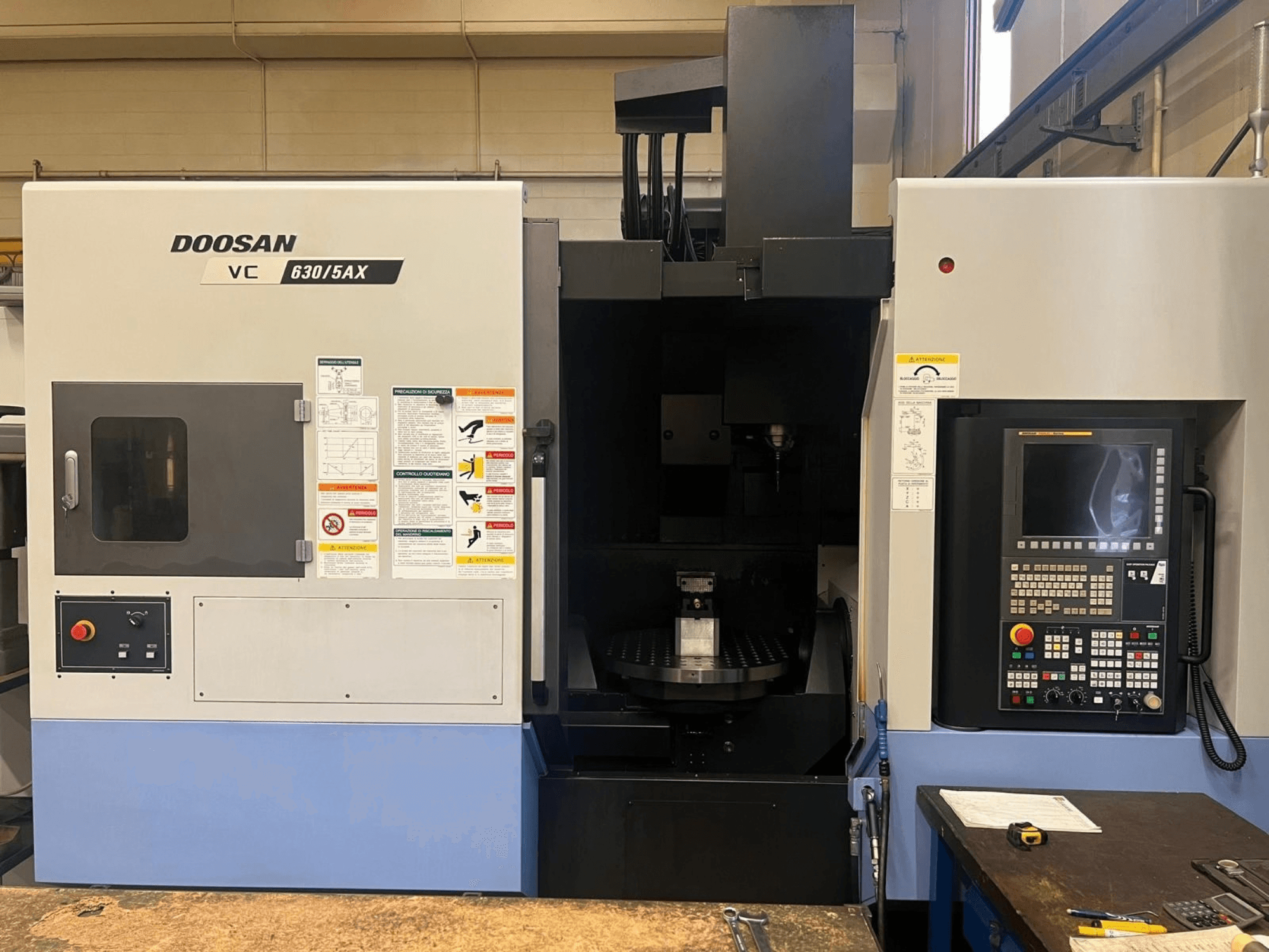 Prednji pogled na vertikalni obradni centar DOOSAN VC 630/5AX, s prikazom upravljačke ploče i područja vretena s vidljivim sigurnosnim naljepnicama.