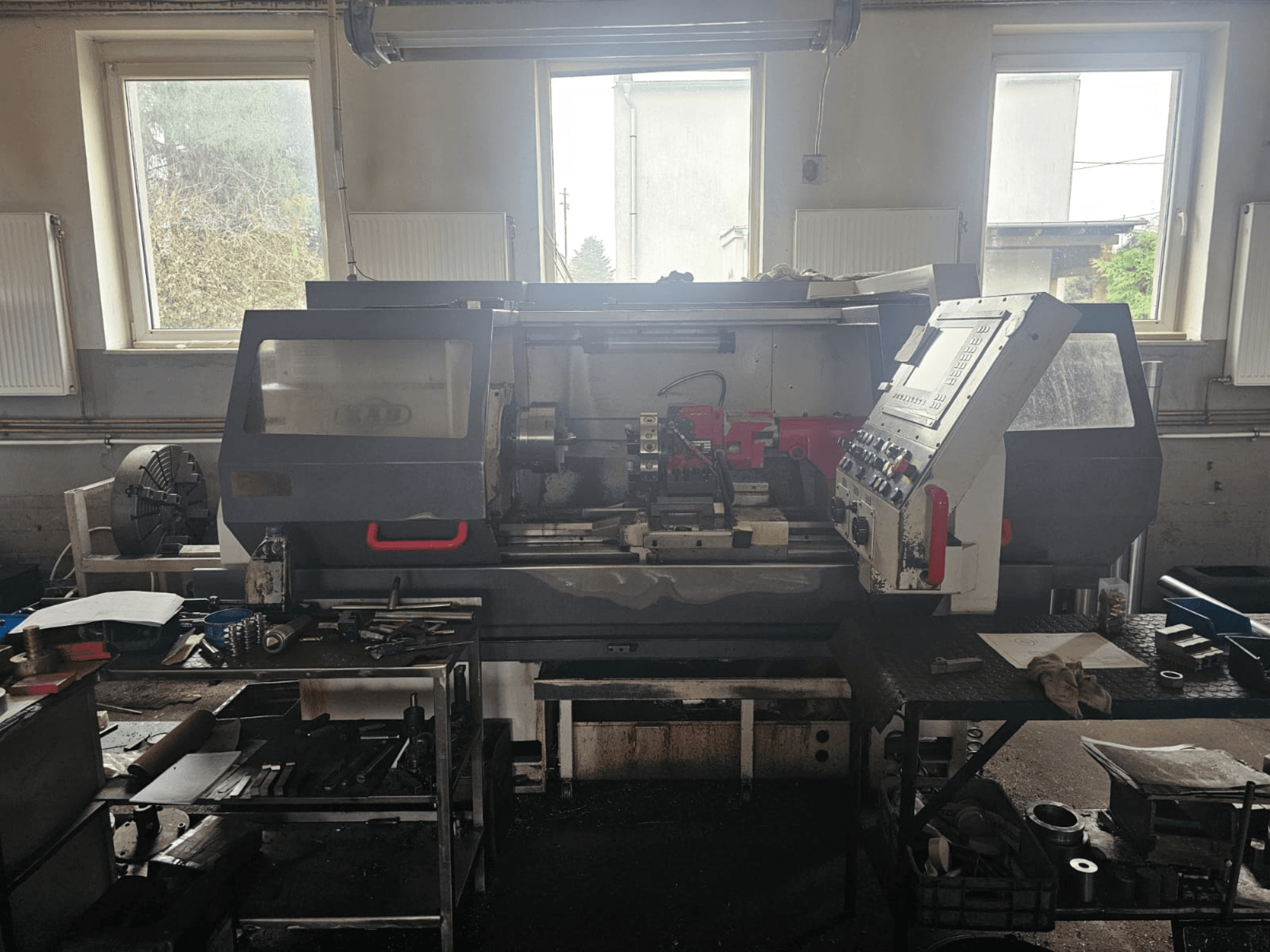 Tokarski stroj u radionici, prednji pogled, s upravljačkom pločom i alatima na stolovima, okružen prozorima i pretrpanim radnim prostorom.