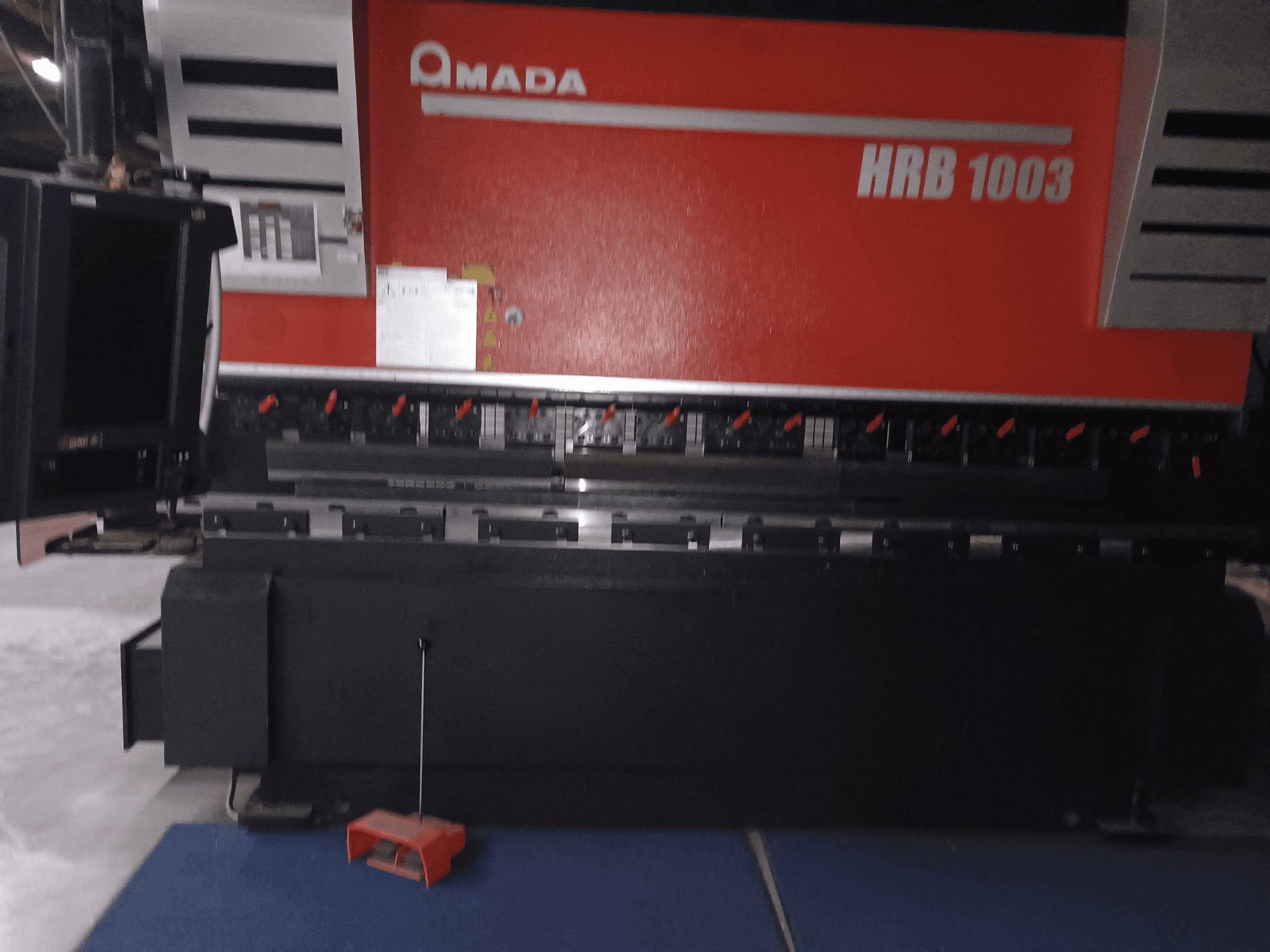 Amada HRB 1003 preša za savijanje, pogled sprijeda; ima upravljačku ploču s crvenim gumbima i crnim kućištem, smještena u industrijskom okruženju.