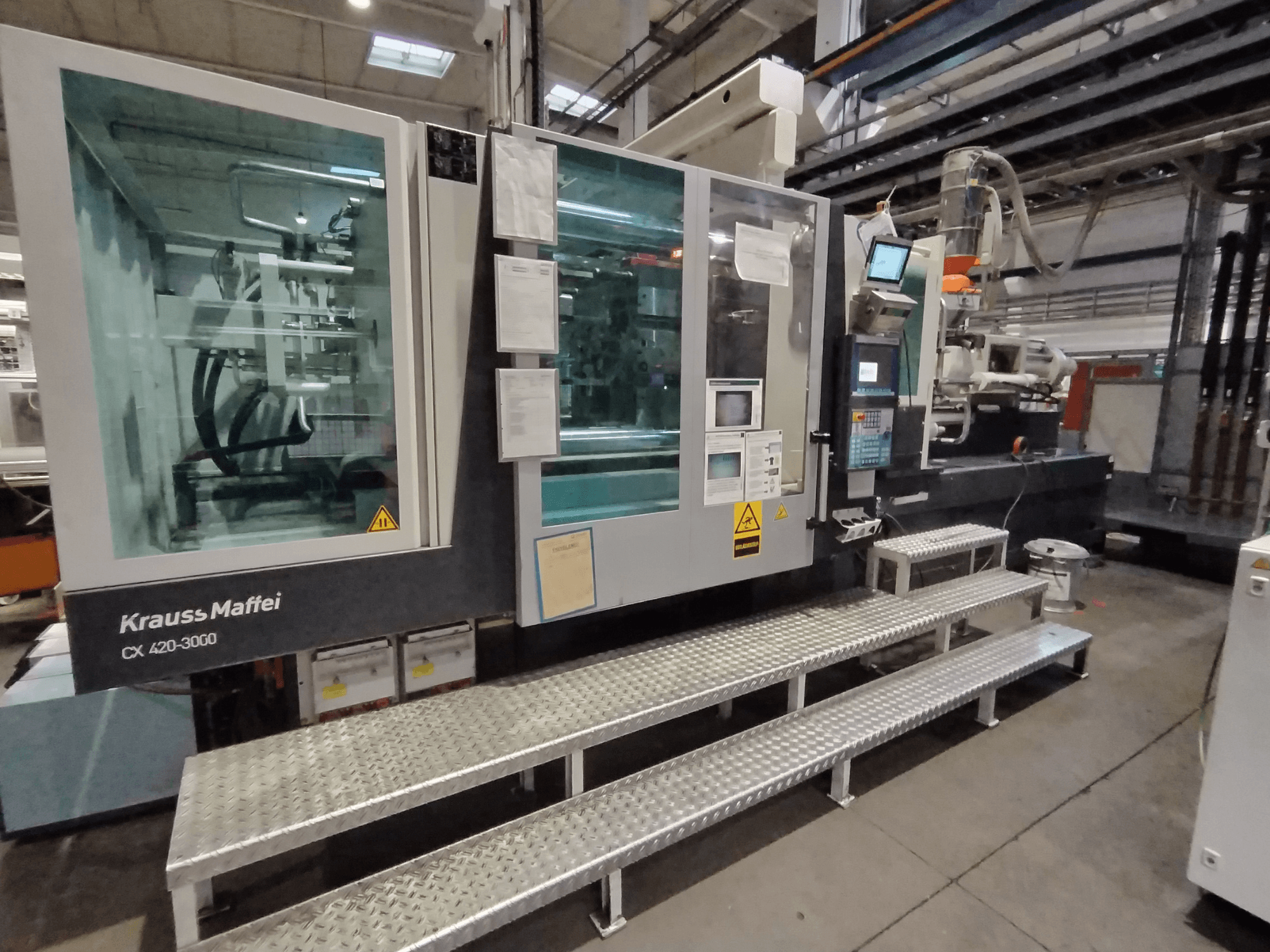 Stroj za brizganje plastike Krauss Maffei CX 420-3000, bočni pogled, s vidljivom upravljačkom pločom i sigurnosnim znakovima, postavljen na metalne stepenice.