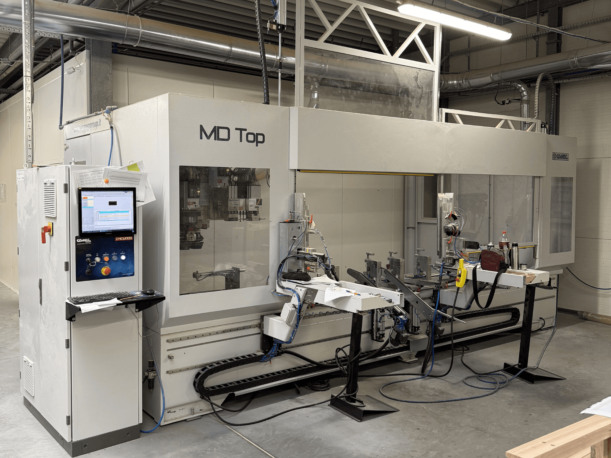 MD Top CNC stroj gledan sprijeda, s upravljačkom pločom, robotskim rukama i radnim prostorom s alatima i materijalima.