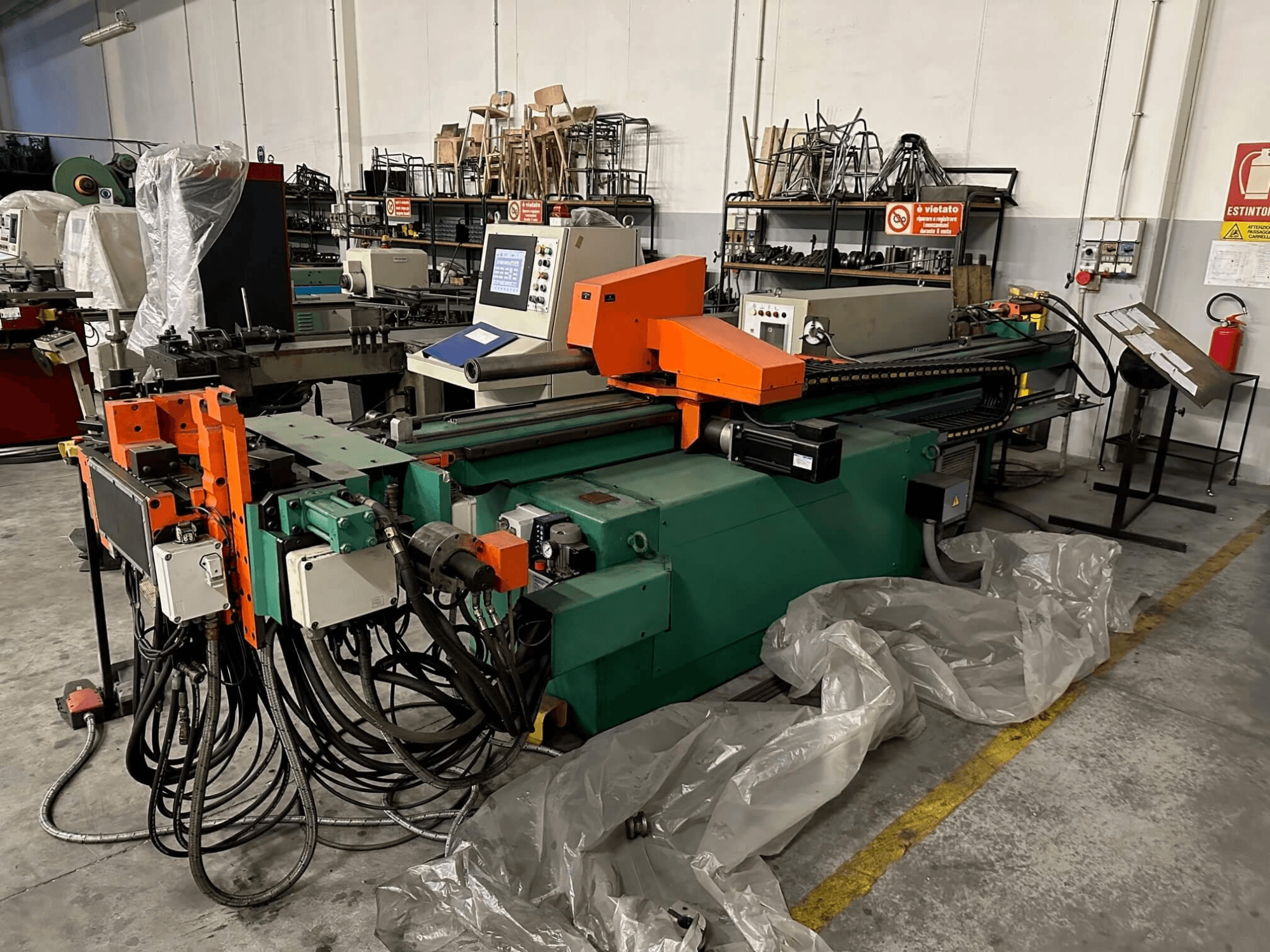 Industrijski stroj s narančasto-zelenim kućištem, upravljačkom pločom i brojnim kabelima, gledan sprijeda u radioničkom okruženju.