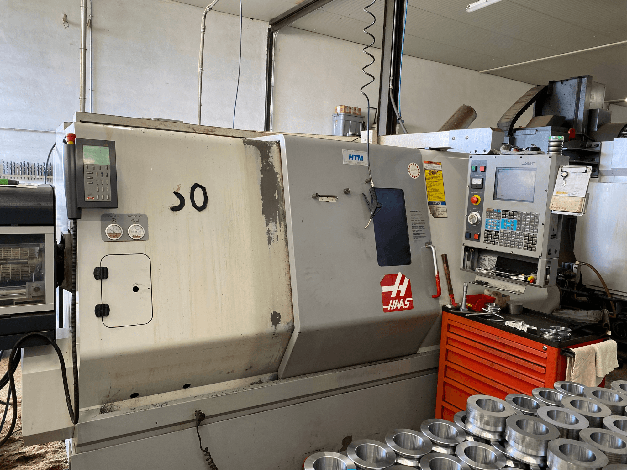 Haas CNC stroj s upravljačkom pločom, prednji pogled, s prikazom tipki za upravljanje, mjerača i metalnih dijelova složenih u blizini.