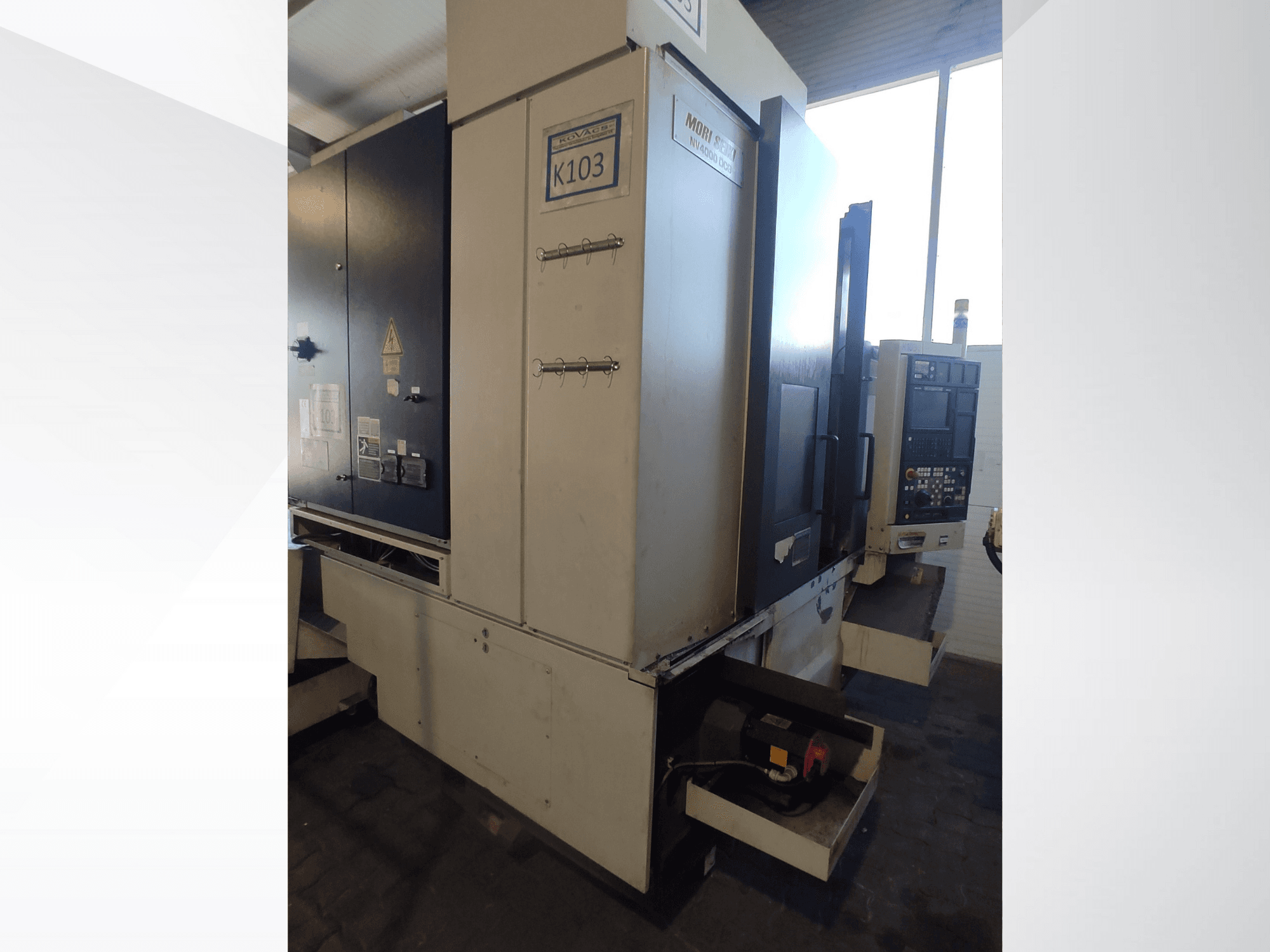 CNC stroj Mori Seiki K103 u prednjem pogledu, s vidljivim upravljačkom pločom, sigurnosnim naljepnicama i električnim komponentama.