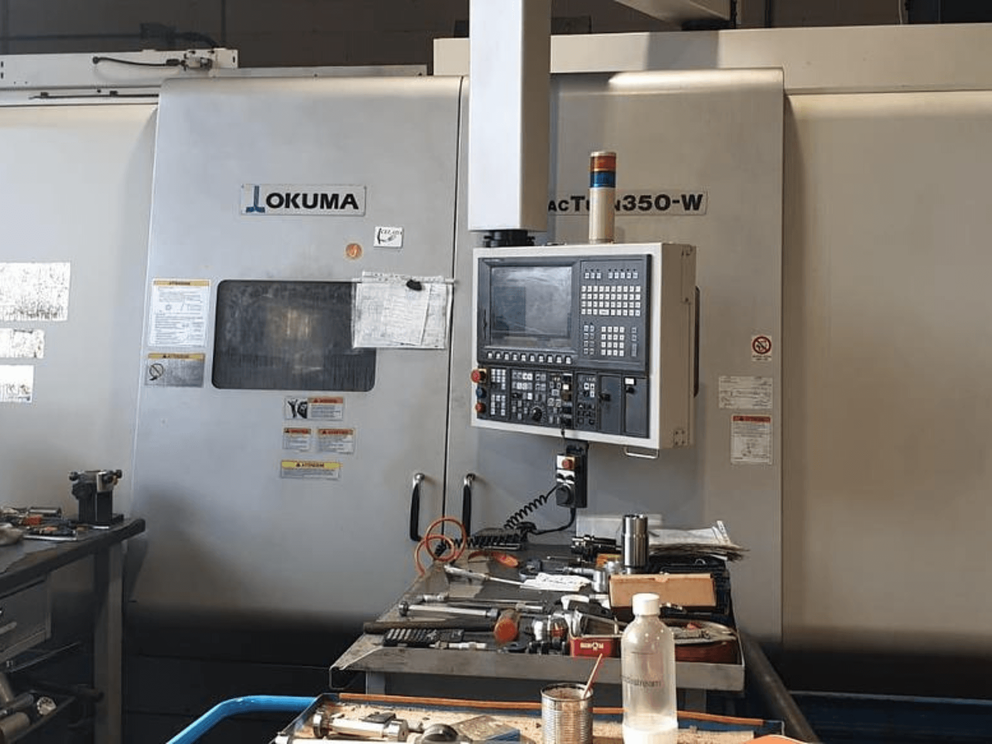 Okuma MAC TURN 350-W tokarsko-glodački centar, prednji pogled s upravljačkom pločom i radnim prostorom zatrpanim alatima i bocom.