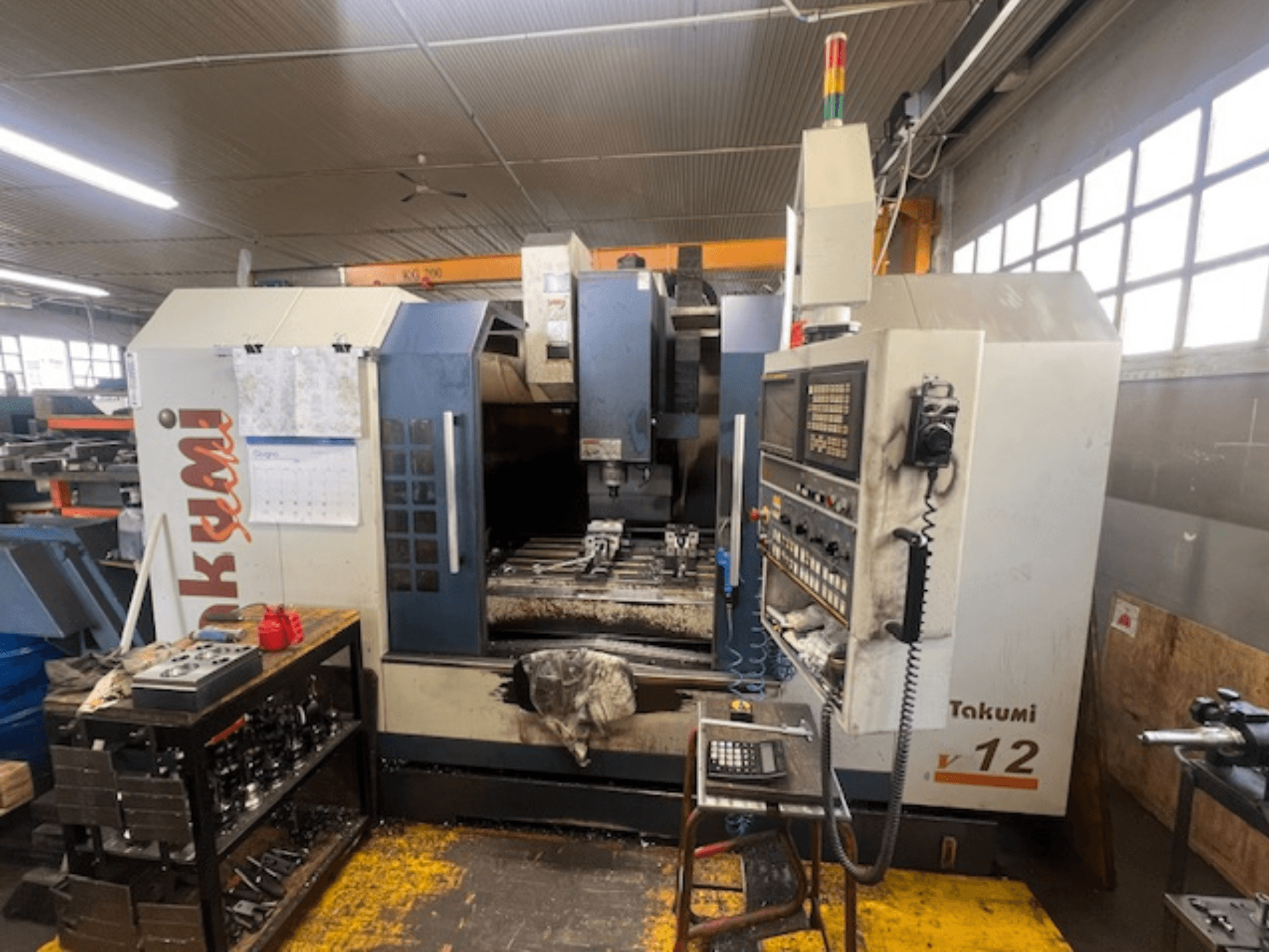 Bočni pogled na CNC glodalicu Takumi V12, koja prikazuje upravljačku ploču, područje rezanja i alate na radnoj stanici.