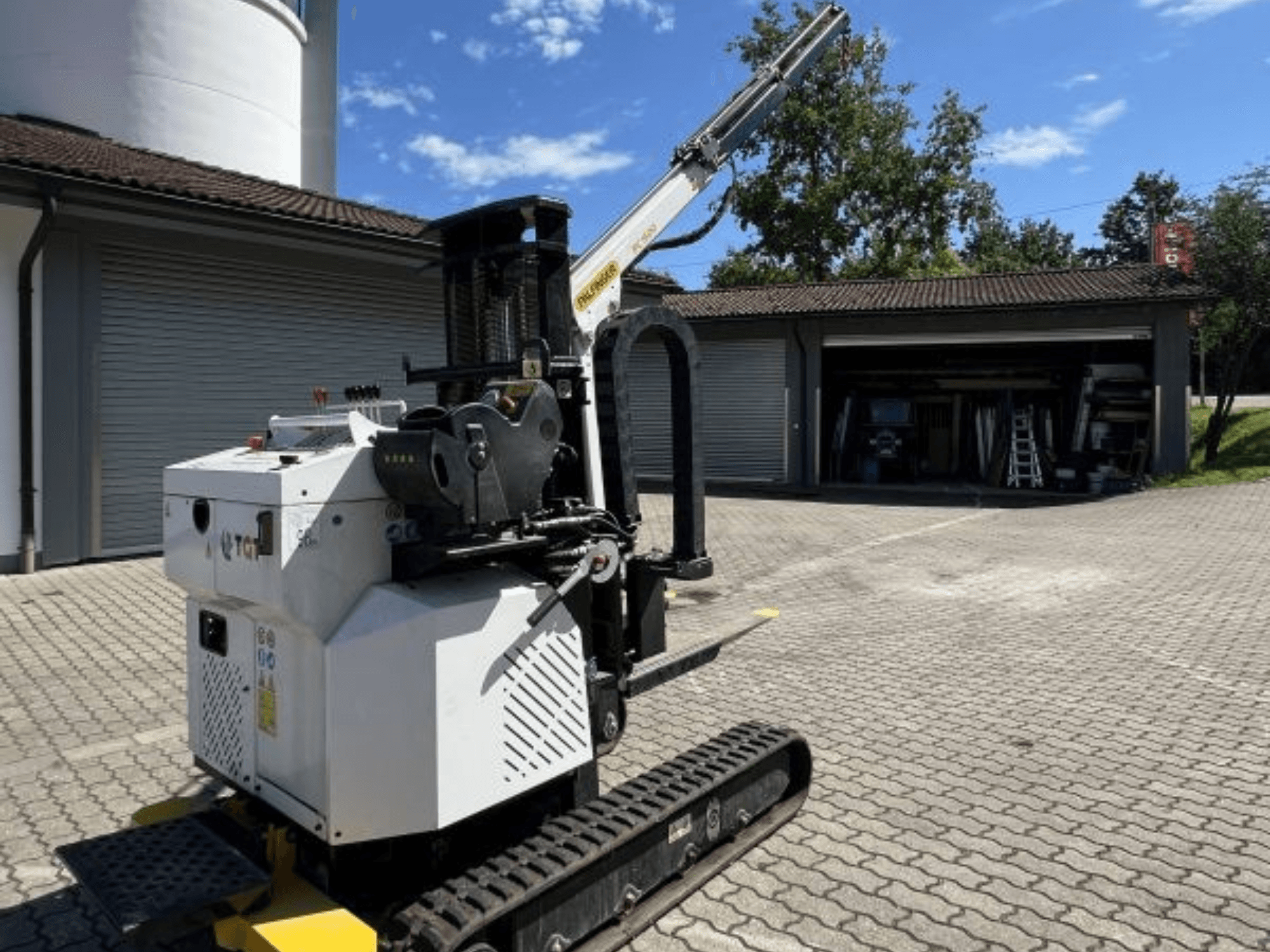 TGI građevinska mini dizalica, bočni pogled, s prikazom gusjenica i montirane ruke, parkirana na ciglenoj površini pored garaže.