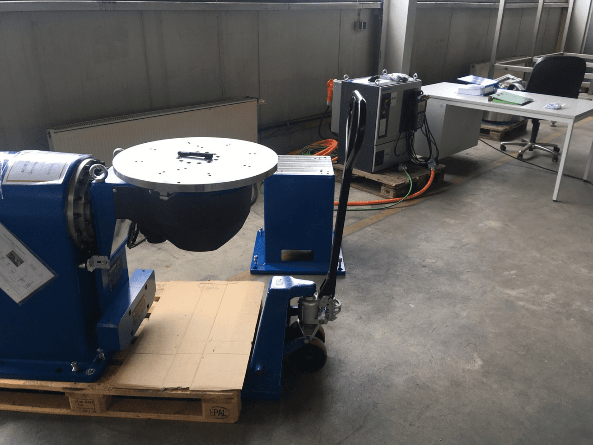 Plava industrijska oprema s kružnom bazom i upravljačkom pločom, gledano sprijeda, na drvenoj paleti u uredskom okruženju.