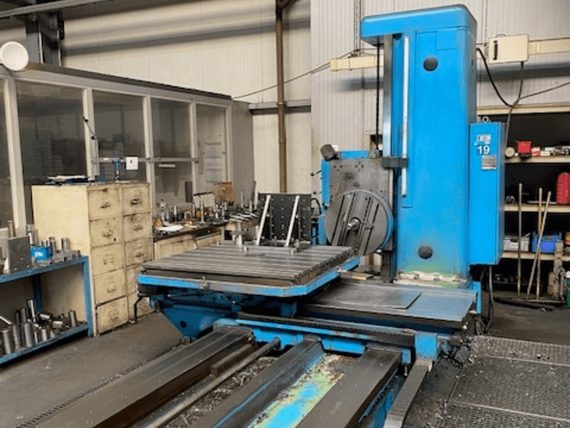 Plava glodalica sprijeda, model broj 19, na podu radionice s alatima i ormarom za pohranu u pozadini.