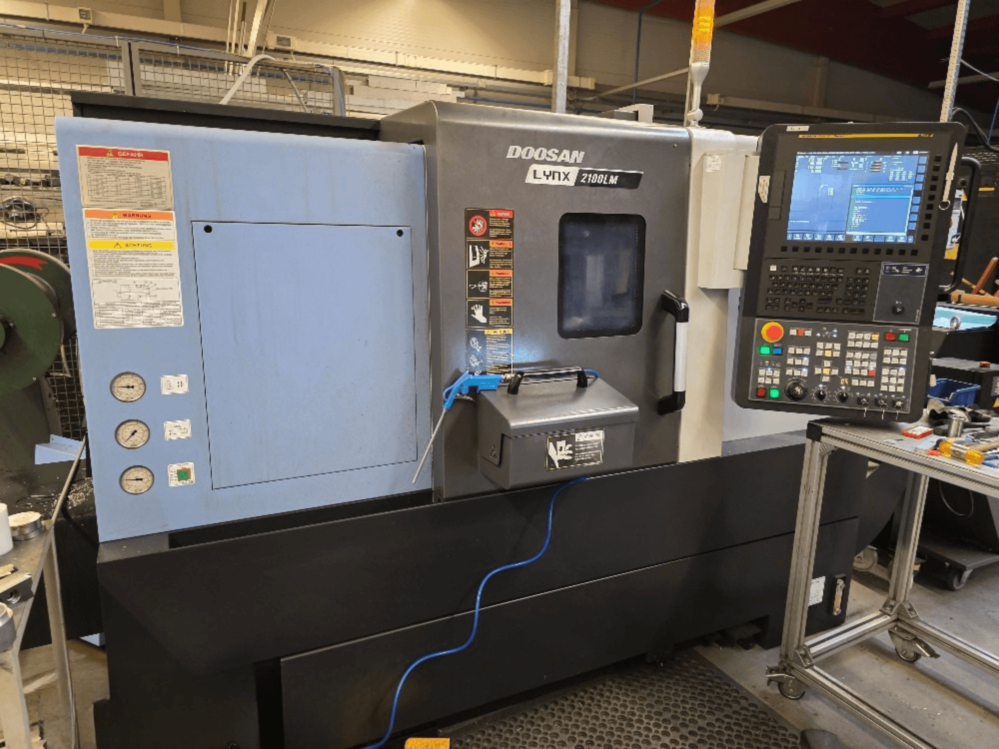 Doosan Lynx 2100LM CNC tokarski stroj, prednji pogled, s upravljačkom pločom, manometrima i sigurnosnim upozorenjima.