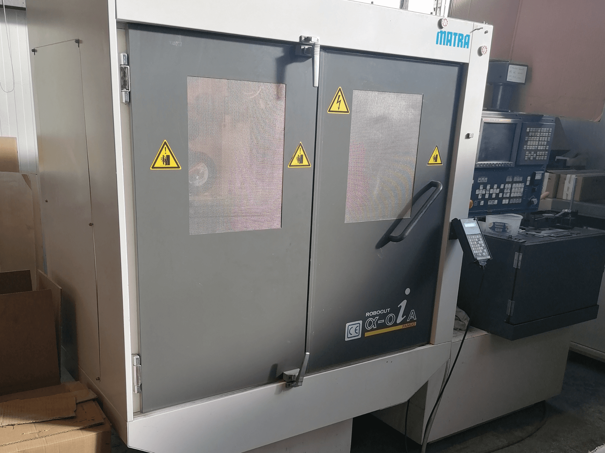 Stroj Robocut α-oZα tvrtke Matra, prednji pogled s prikazom sigurnosnih upozorenja, upravljačke ploče i dvostrukih vrata s mrežastim prozorima.