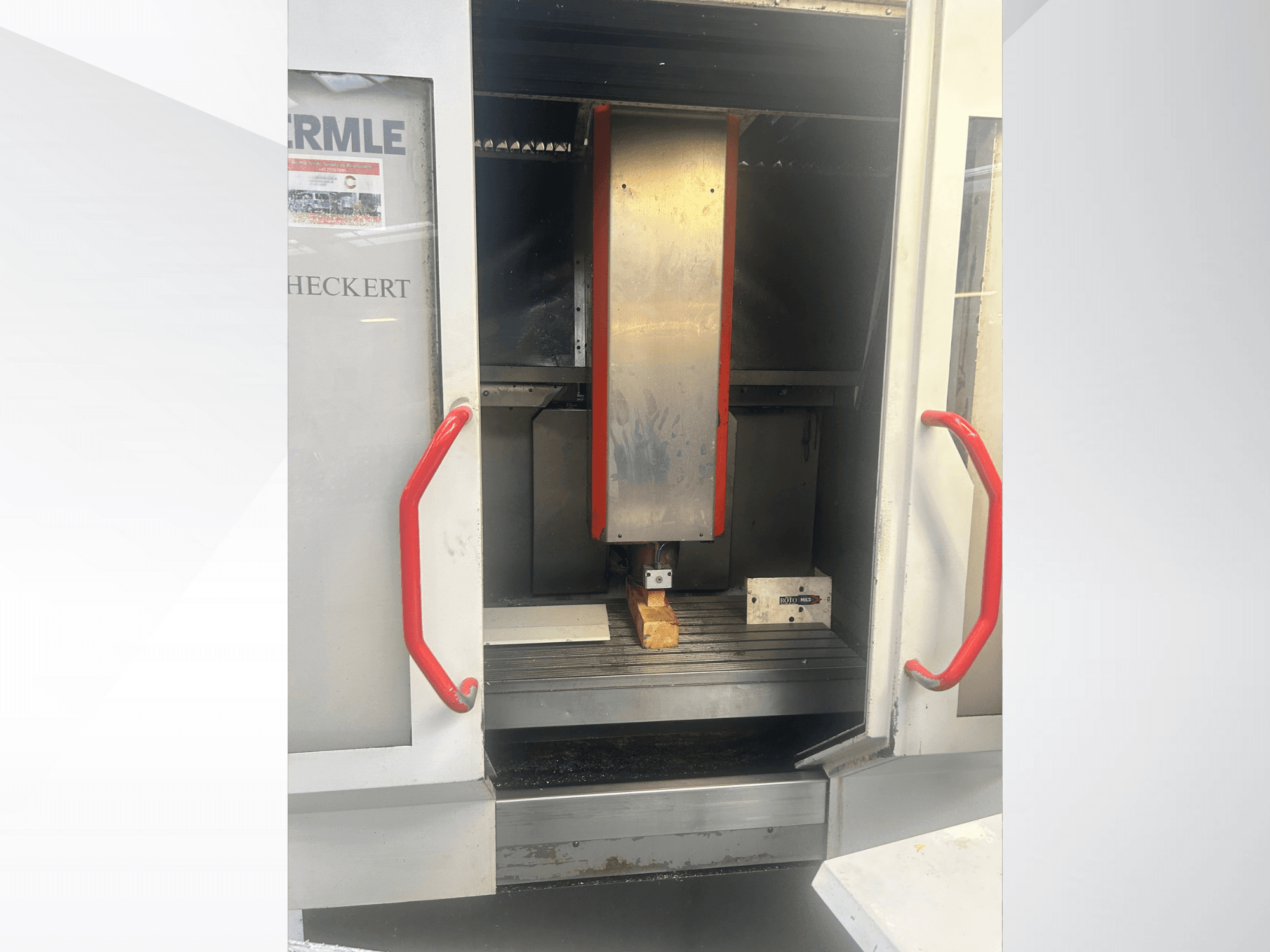 Prednji pogled na Hermle CNC stroj s metalnom unutrašnjosti, crvenim ručkama i radnim komadom na stolu.