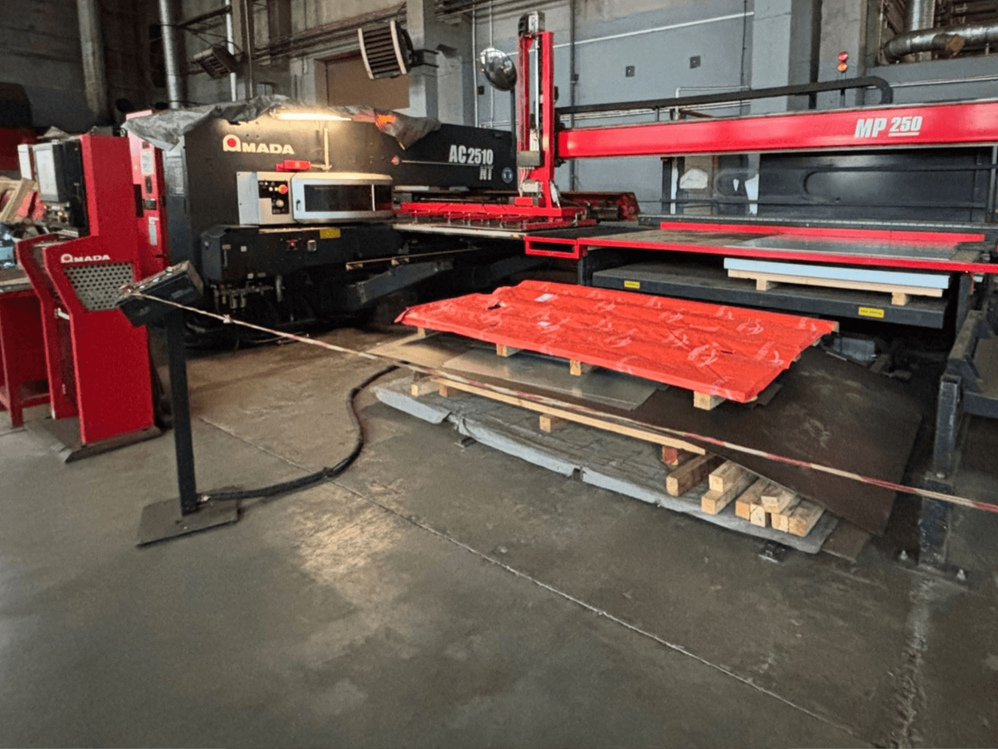 Amada AC2510 NT laserski rezač u tvorničkom okruženju, prednji pogled, teški strojevi s metalnim pločama i paletama na podu.