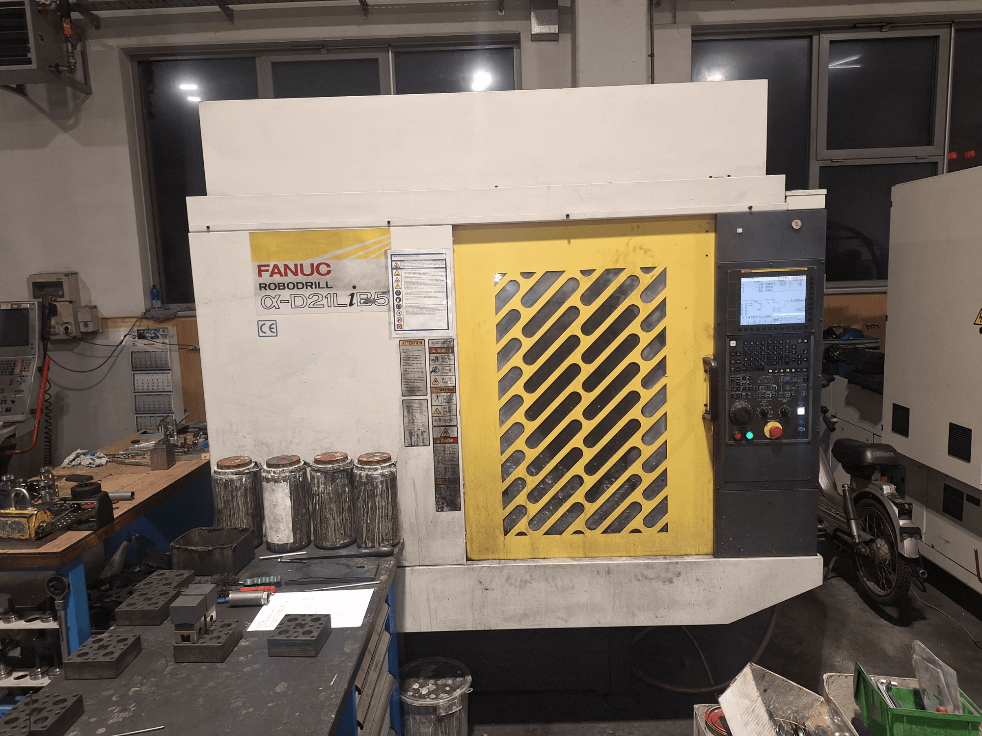 Prednji pogled na FANUC Robodrill α-D21LiB5, s upravljačkom pločom, žutom rešetkom i obližnjim alatima na radnom stolu.
