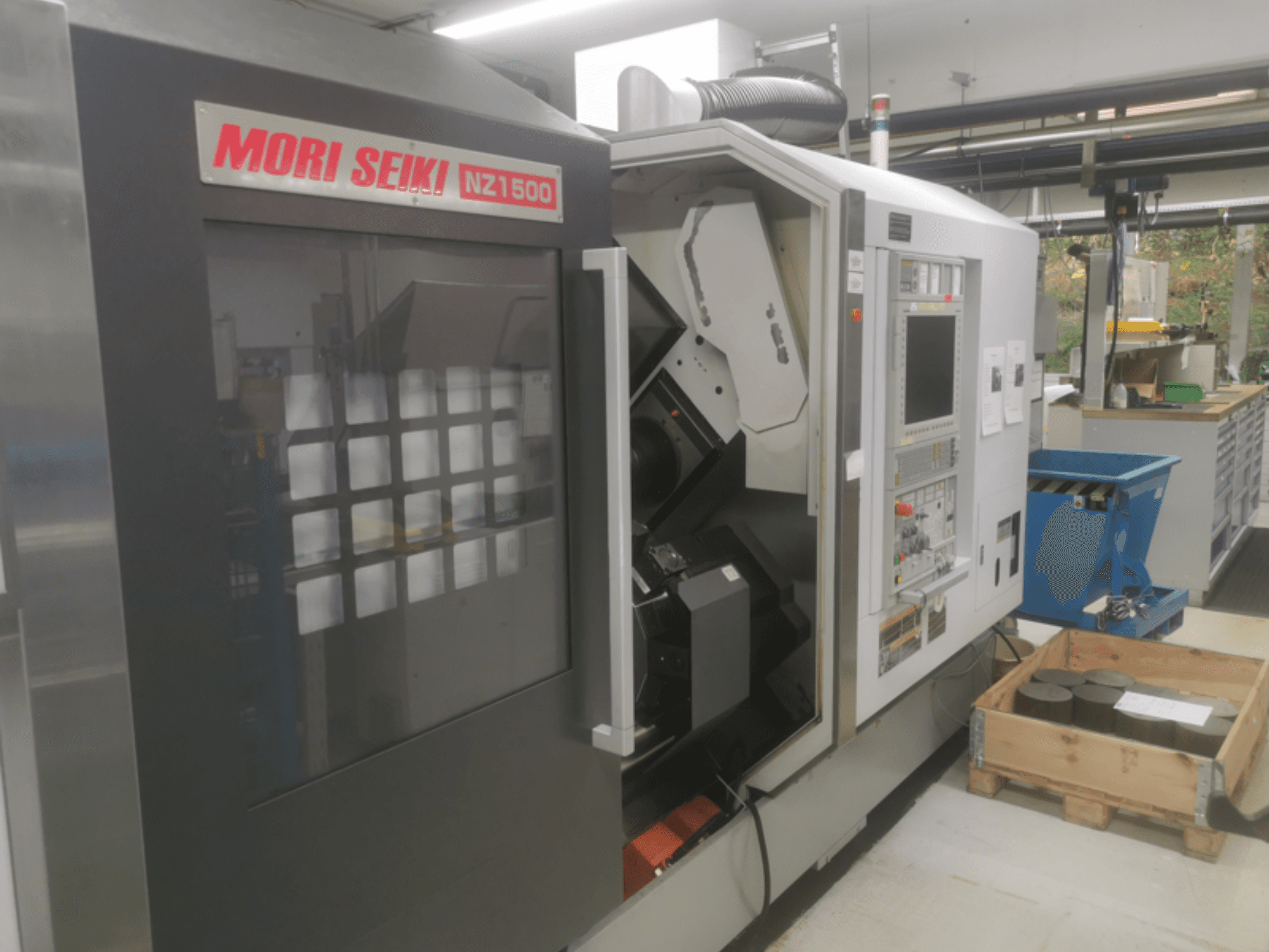 MORI SEIKI NZ1500 CNC stroj pod blagim kutom, prikazuje upravljačku ploču i radni prostor s metalnim obratcima na drvenoj paleti.