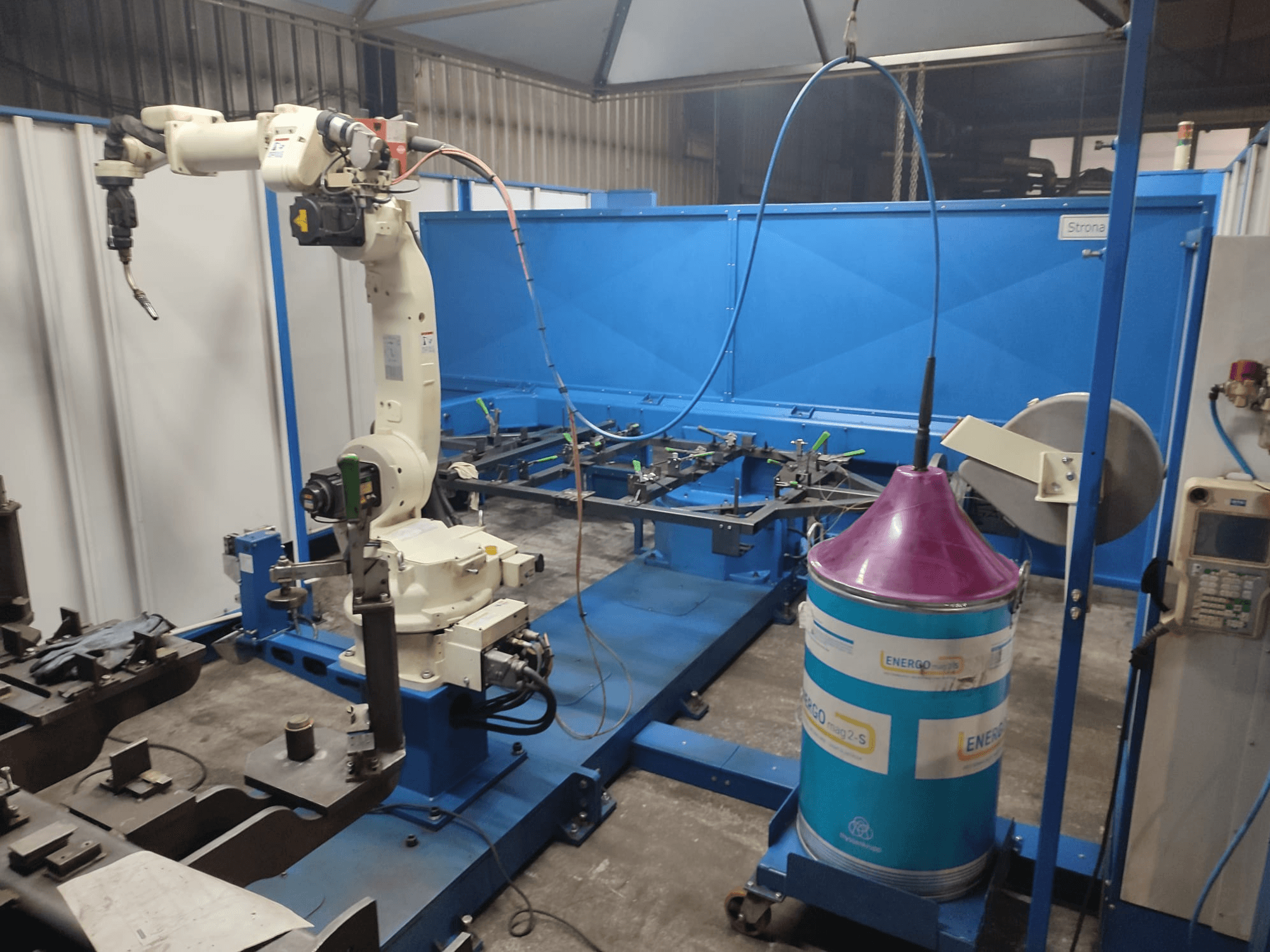 Industrijska robotska ruka s bijelo-plavom bazom, postavljena u radionici s plavom radnom površinom i metalnim komponentama.