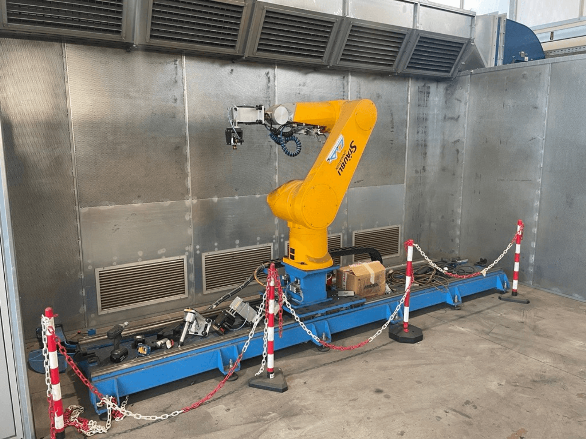 STAUBLI CS8C-RX160 robotska ruka u radionici, bočni pogled, s kabelima i raznim alatima na plavoj podlozi okruženoj zaštitnim barijerama.