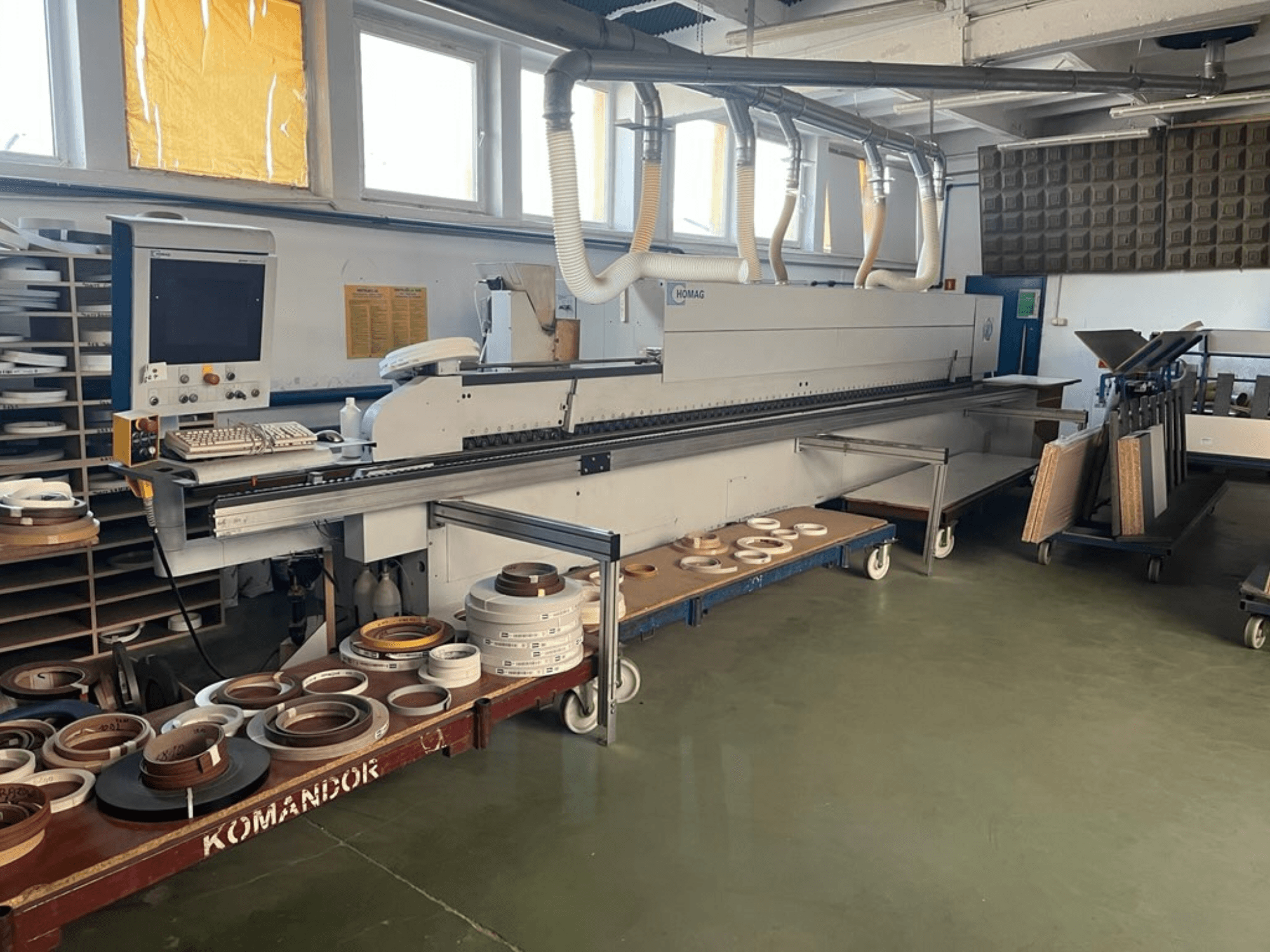 HOMAG stroj za rubno lijepljenje u radionici, s upravljačkim elementima i raznim materijalima za rubno lijepljenje na plavim kolicima. Bočni pogled.