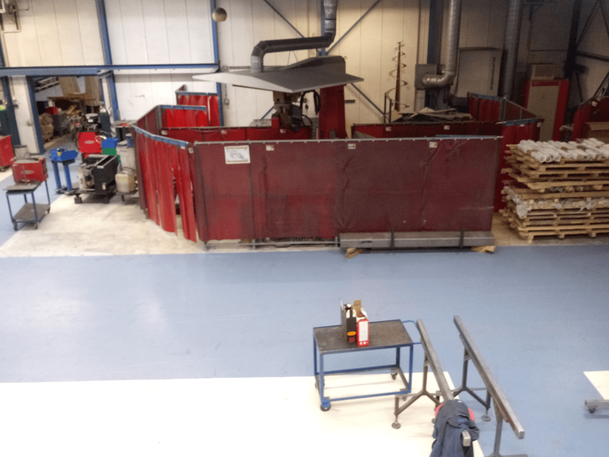 Industrijski radni prostor s crvenim zavjesama za zavarivanje, strojevima i paletama; pogled odozgo koji prikazuje raspored i opremu.