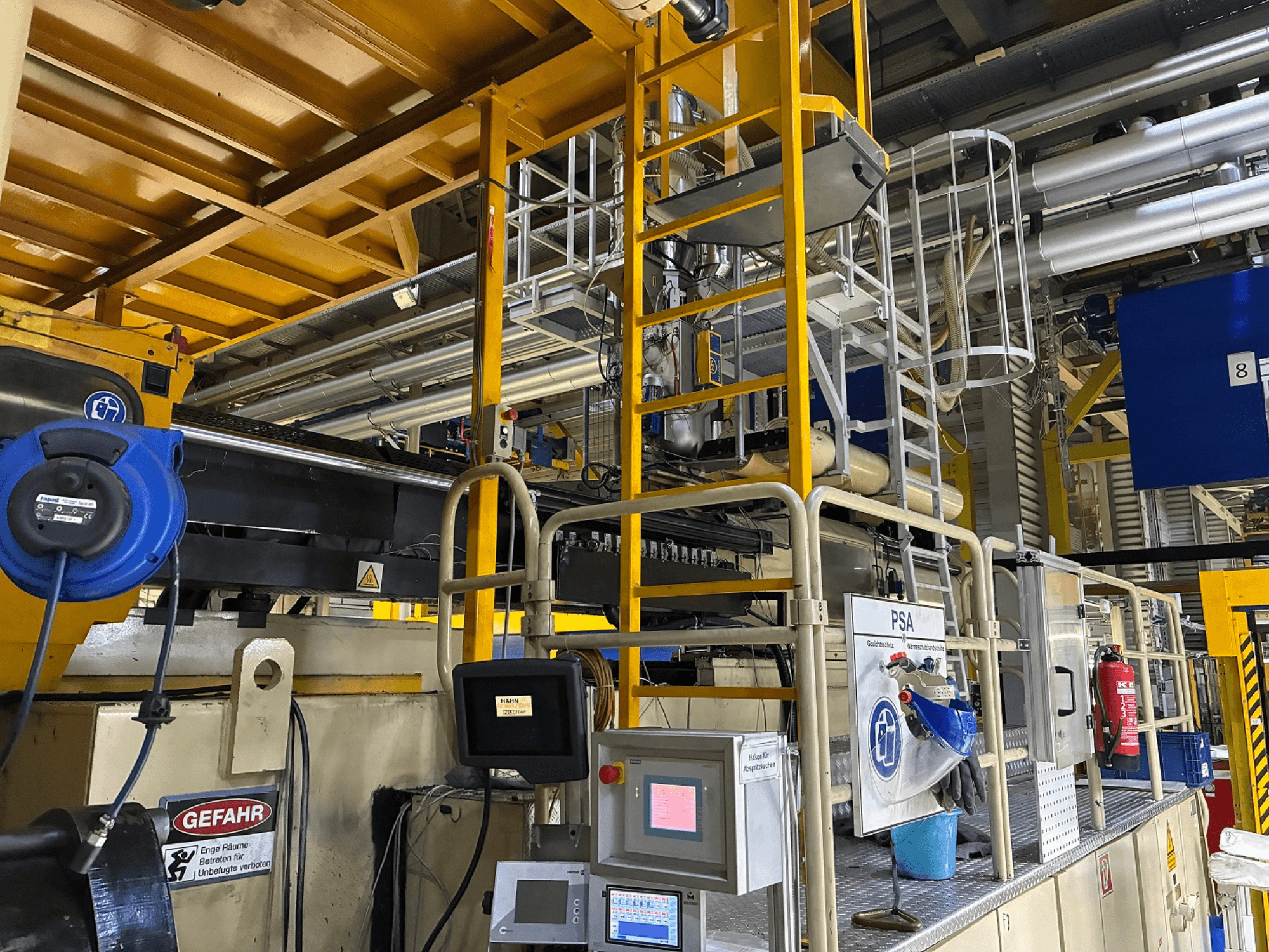 Upravljačka ploča industrijskih strojeva s plavom sigurnosnom opremom i žutim skelama, bočni pogled s cijevima i znakovima upozorenja.