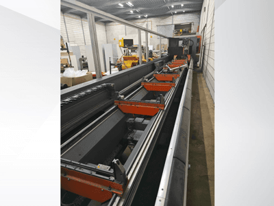 Bočni pogled na industrijski CNC stroj s linearnim vodilicama, narančastim stezaljkama i preciznim komponentama u skladišnom okruženju.