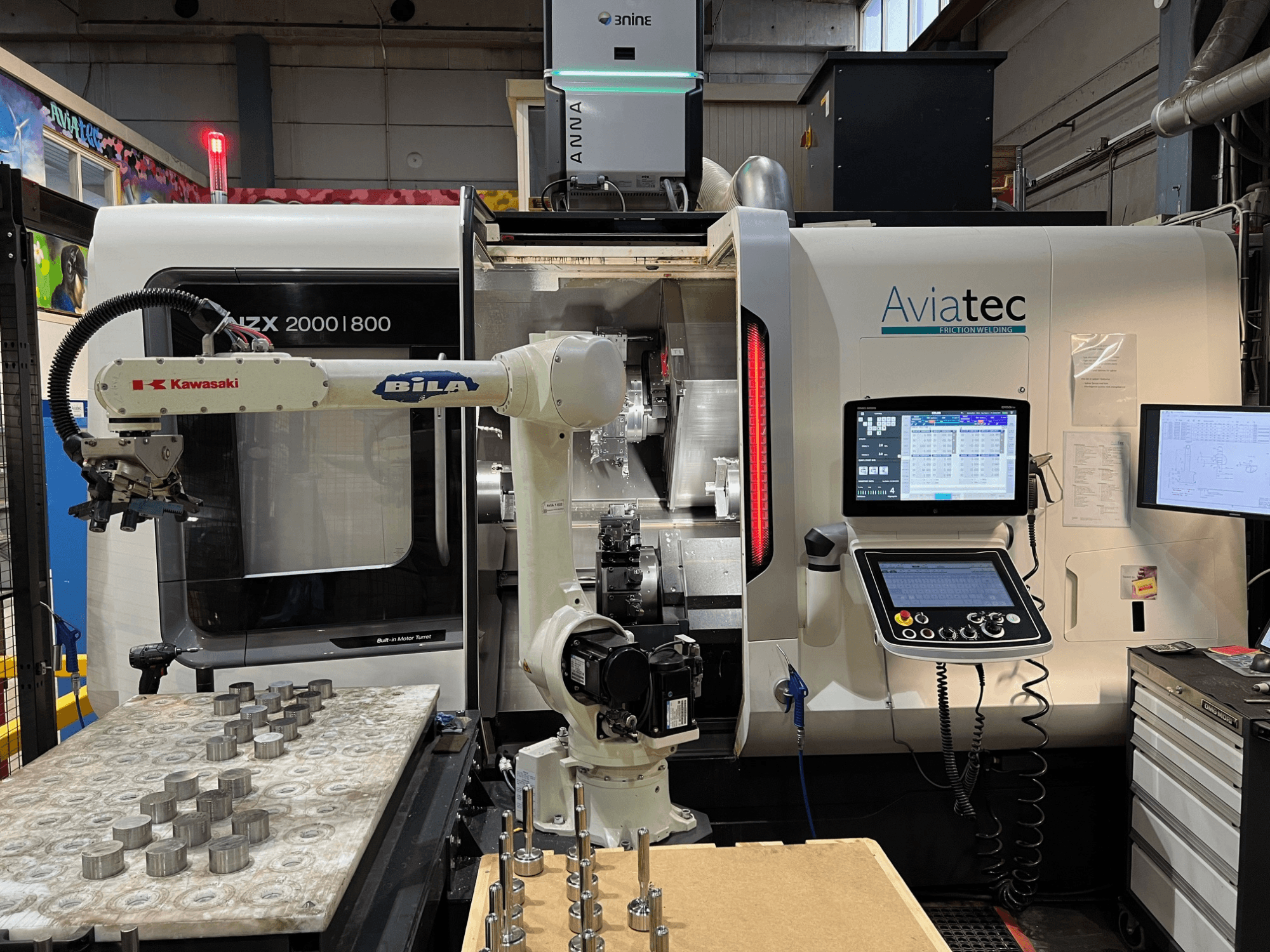 Horizontalni tokarski stroj DMG MORI NZX 2000/800SY s Kawasaki robotskom rukom, upravljačkom pločom i metalnim komponentama na radnom stolu.