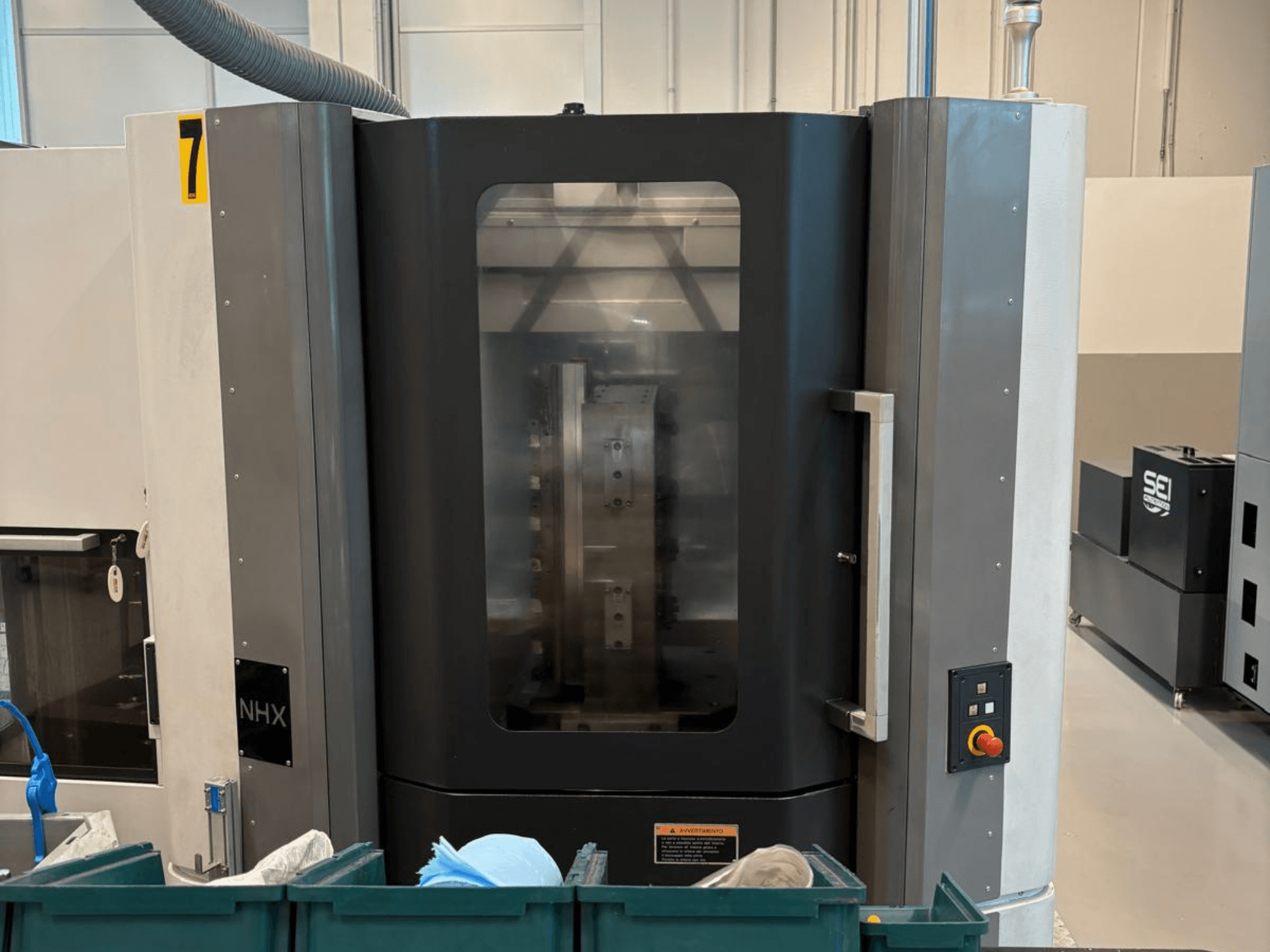 NHX CNC stroj u prednjem pogledu, s upravljačkom pločom i jasnim zaslonom; smješten u radionici sa zelenim kantama za smeće u blizini.