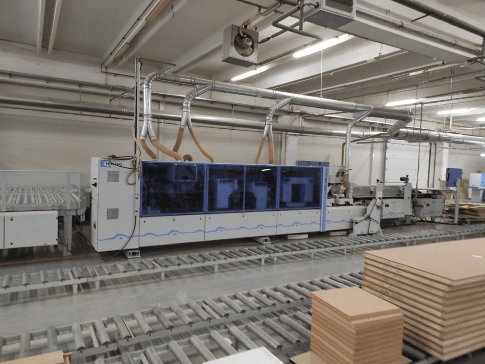 HOMAG proizvodni stroj u tvornici, pogled sprijeda, s plavim kućištem i transportnim sustavom sa složenim listovima šperploče.