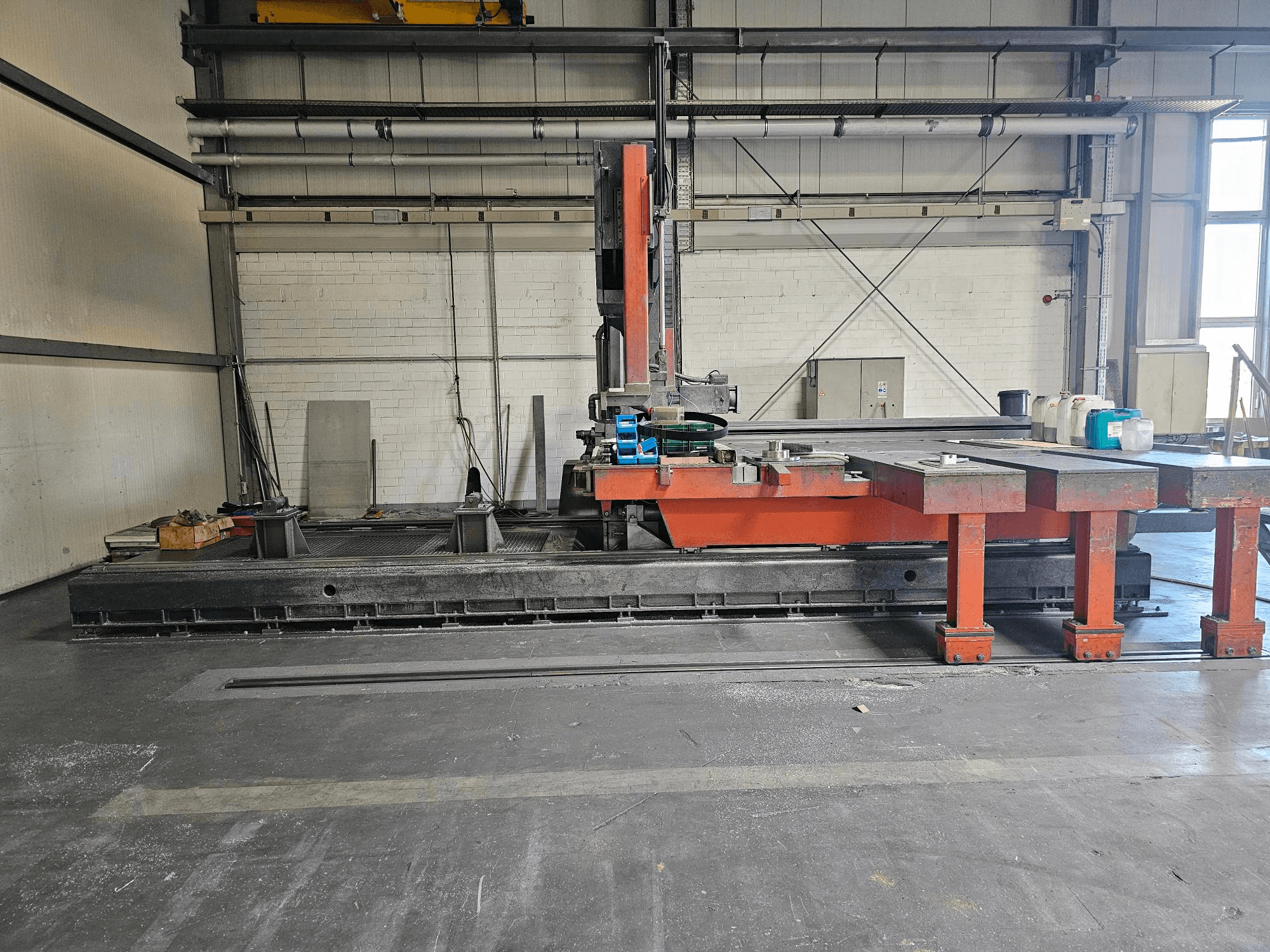 Industrijski stroj za rezanje u otvorenoj radionici, narančaste i crne boje s velikim stolom; bočni pogled koji prikazuje pribor i radni prostor.