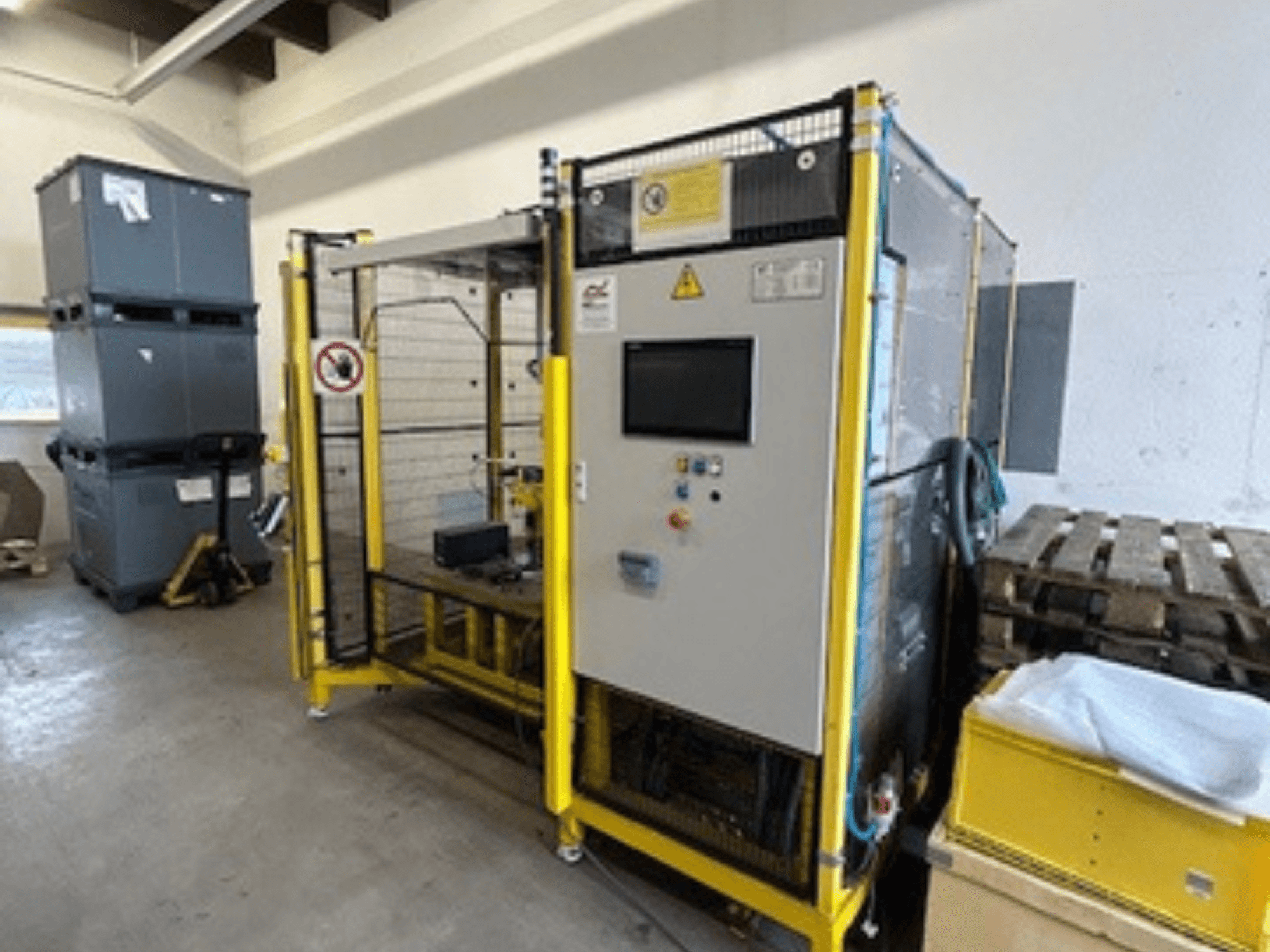 Industrijski stroj s upravljačkom pločom i sigurnosnim značajkama, prednji pogled; žuti okvir, sive kante i palete u pozadini.