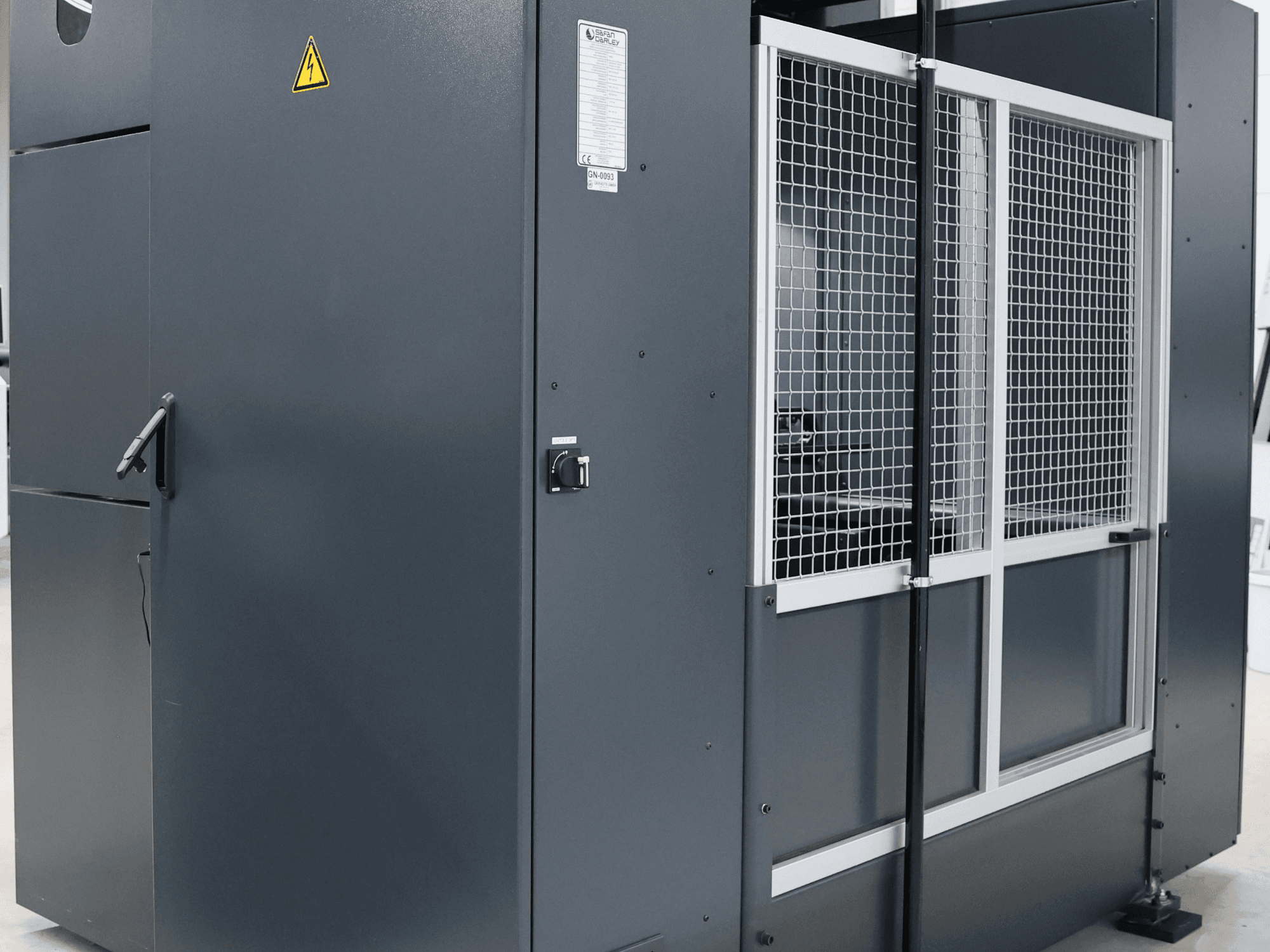 Sivi industrijski stroj s upozorenjem o sigurnosti, prednji pogled s mrežastim vratima i upravljačkom pločom, označen model.