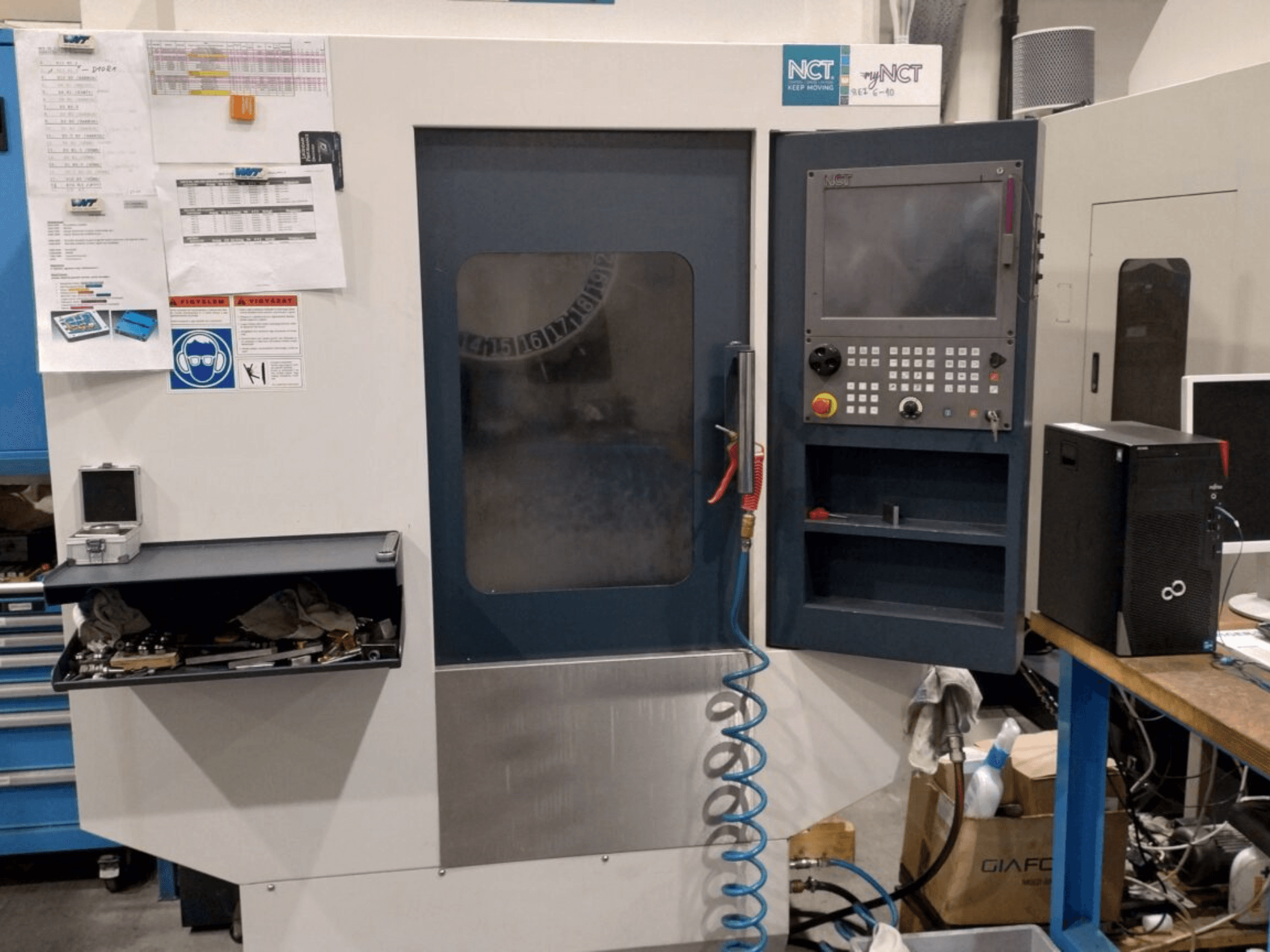 NCT CNC stroj, prednji pogled, sadrži upravljačku ploču s numeričkom tipkovnicom, alate u ladici za pohranu i priručnik na zidu.