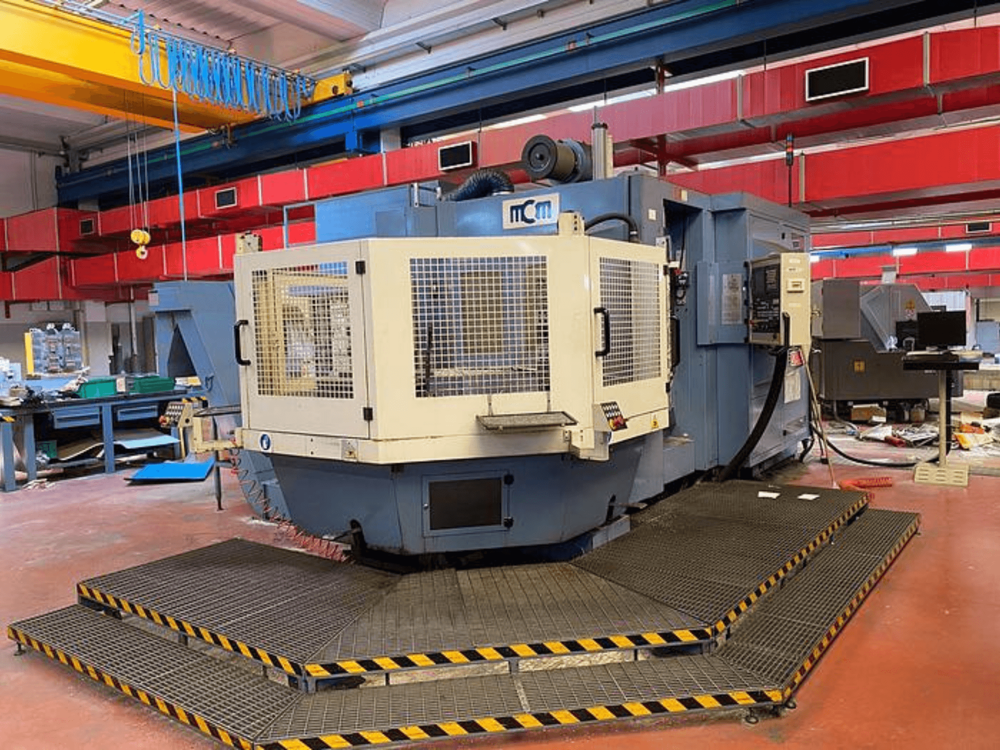 MCM CNC stroj u tvornici, prednji pogled sa zaštitnim kavezom, crnim i žutim sigurnosnim rubovima, okružen industrijskom opremom.