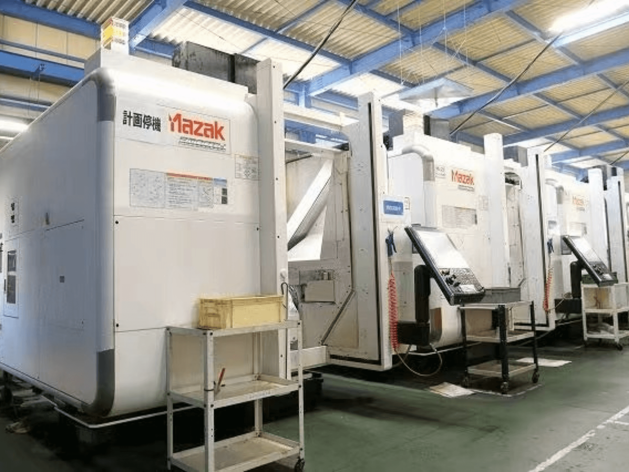 Mazakovi CNC strojevi u tvorničkom okruženju, bočni pogled s upravljačkim pločama i naljepnicama na bijelim kućištima.