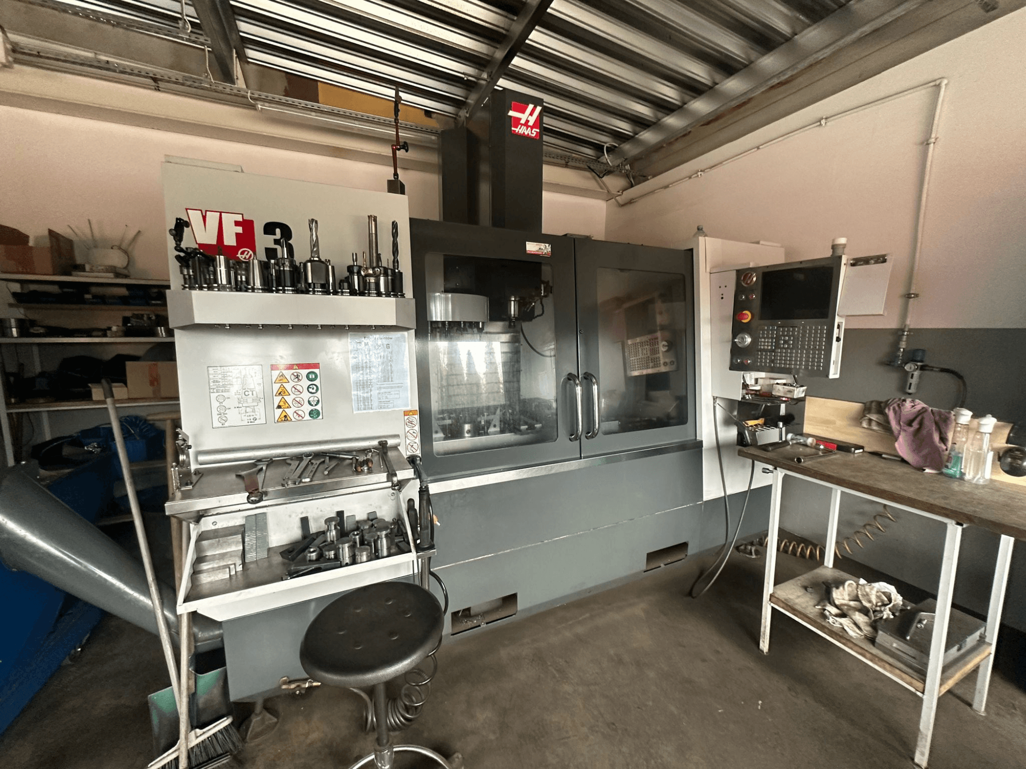 Haas VF-3 CNC stroj u radionici, prednji pogled s alatima i kontrolama na susjednom stolu, prikazuje tehničko sučelje i sigurnosne informacije.