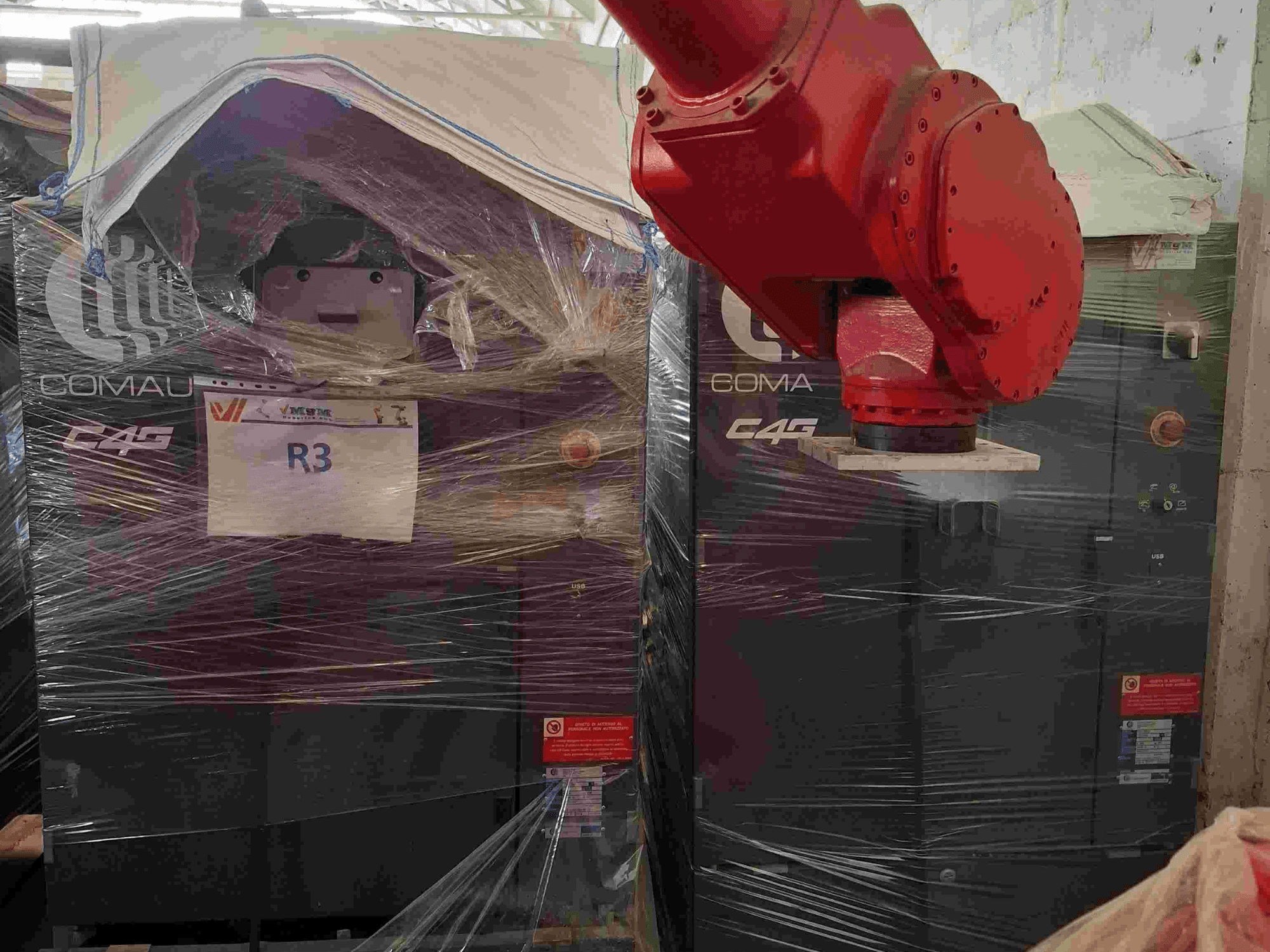 Robotska ruka Comau C4G prikazana sprijeda, omotana plastikom, označena s "R3", s vidljivim naljepnicama upozorenja na stroju.