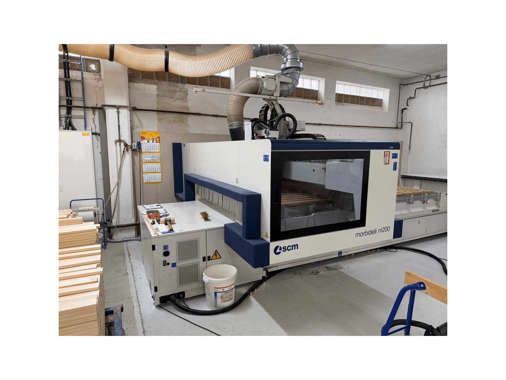 CNC obradni centar SCM Morbidelli M200F u radionici, bočni pogled s drvenim komadima na stolu i ventilacijskim sustavom iznad.
