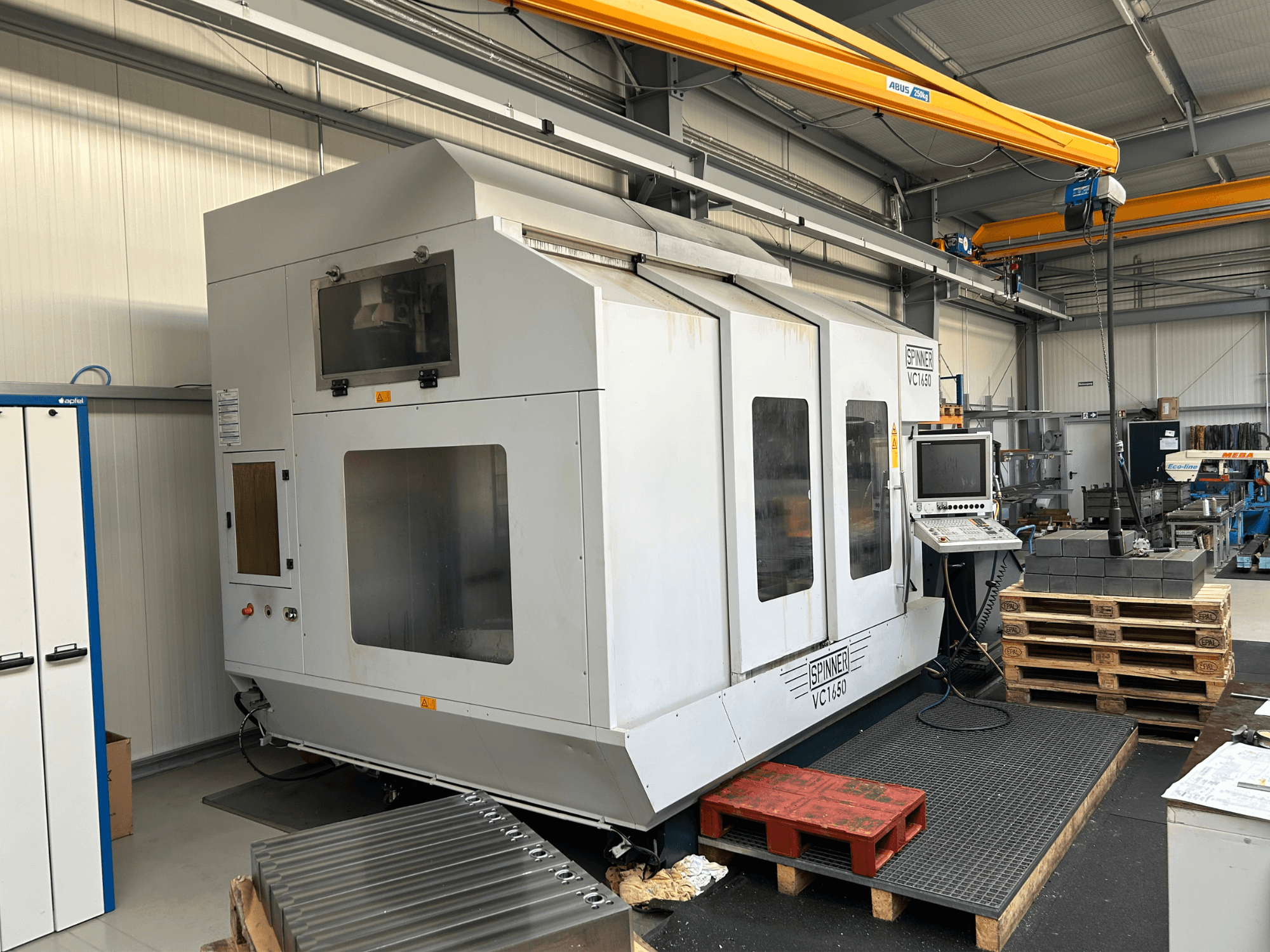 CNC stroj Spinner VC 1650, pogled sprijeda, s upravljačkom pločom, velikim prozorima i paletama naslaganim u blizini u radioničkom okruženju.